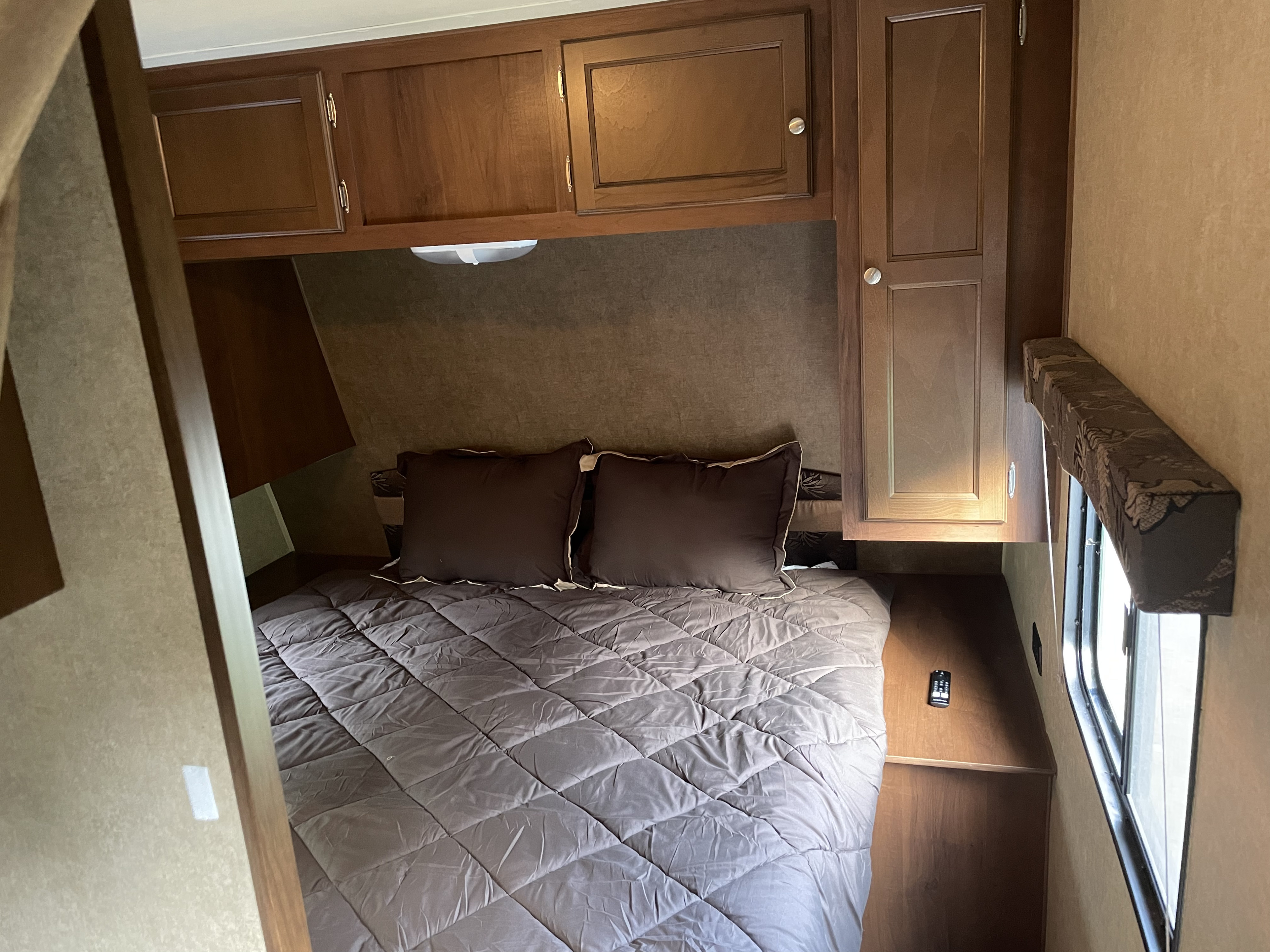 Cozy RV rental bedroom with a comfortable bed, dark bedding, overhead wooden cabinets, and a side window.. Jayco Jay Flight Swift SLX 2014