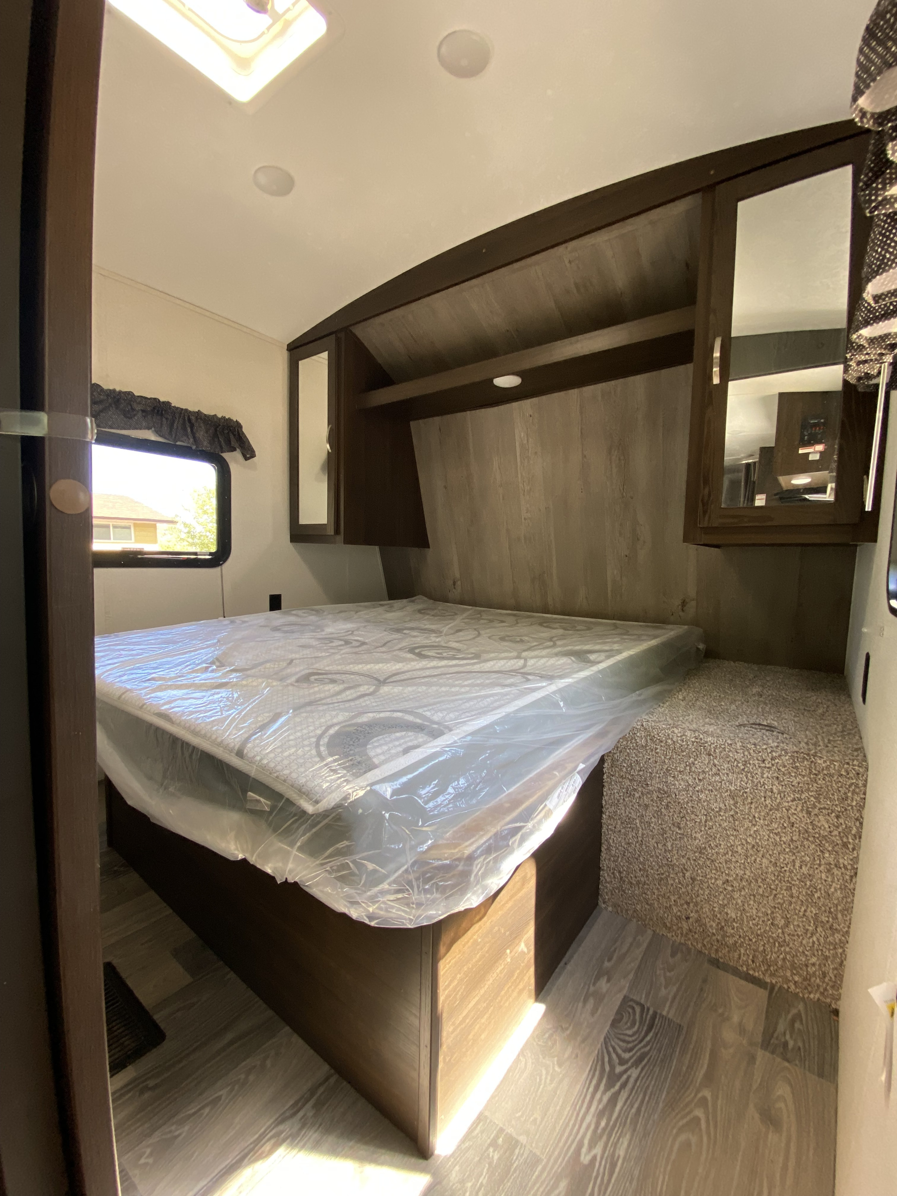 RV rental bedroom: a bed with plastic-wrapped mattress, overhead cabinets, window, and wood-look/carpet flooring.. Keystone RV Springdale 2021