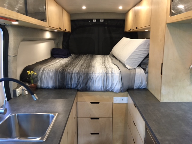 This rv rental features a cozy bed, kitchen sink, and wood cabinets, offering comfort in a compact van interior.. Ford Transit 2015