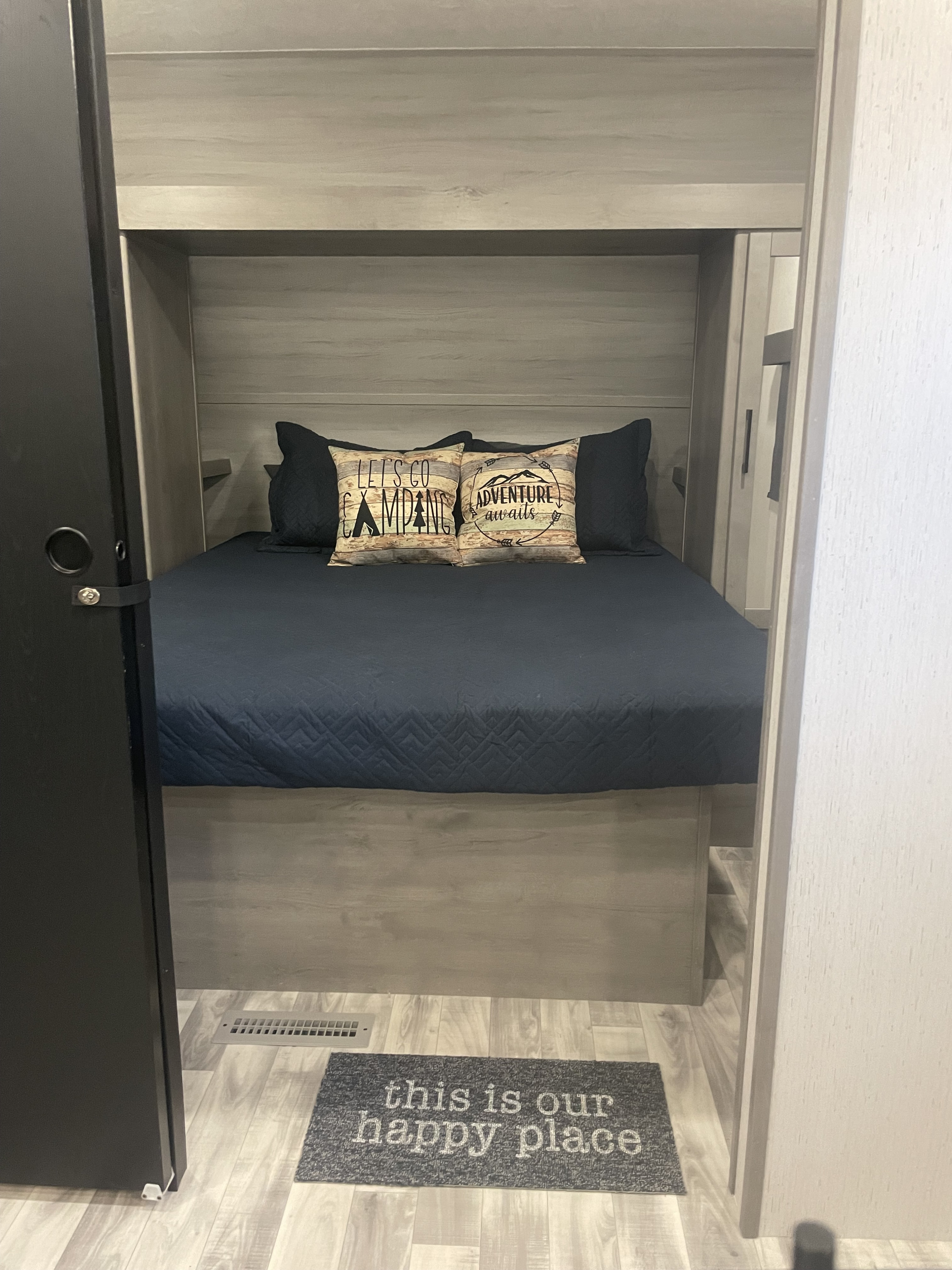 View of a compact RV rental bedroom with a dark blue bed, two camping-themed pillows, and a "happy place" floor mat.. Dutchman Kodiak SE 2022