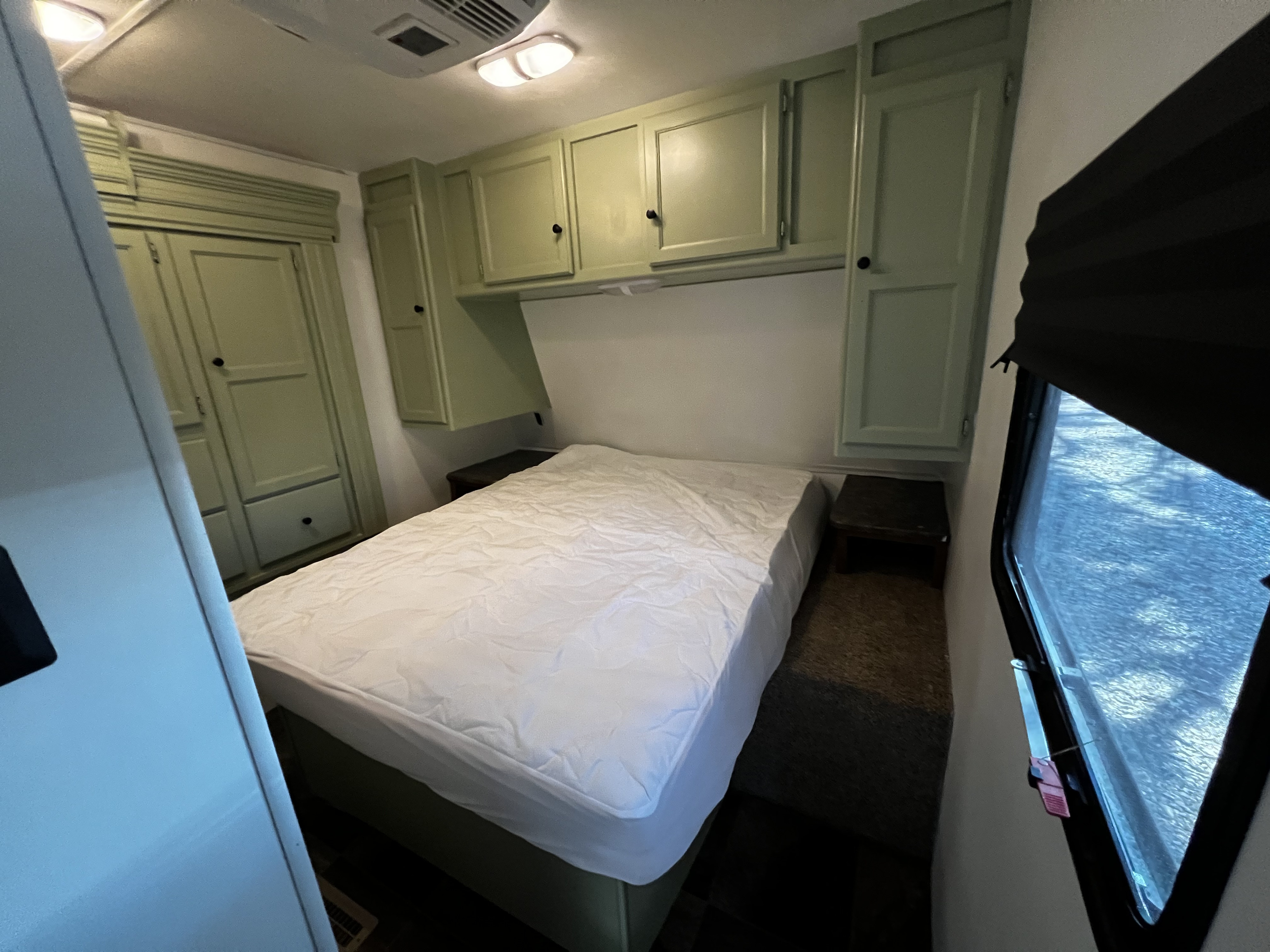 RV rental bedroom showing a bed with white mattress pad, light green overhead cabinets, and a side wardrobe, plus a window.. Crossroads Rezerve 2016