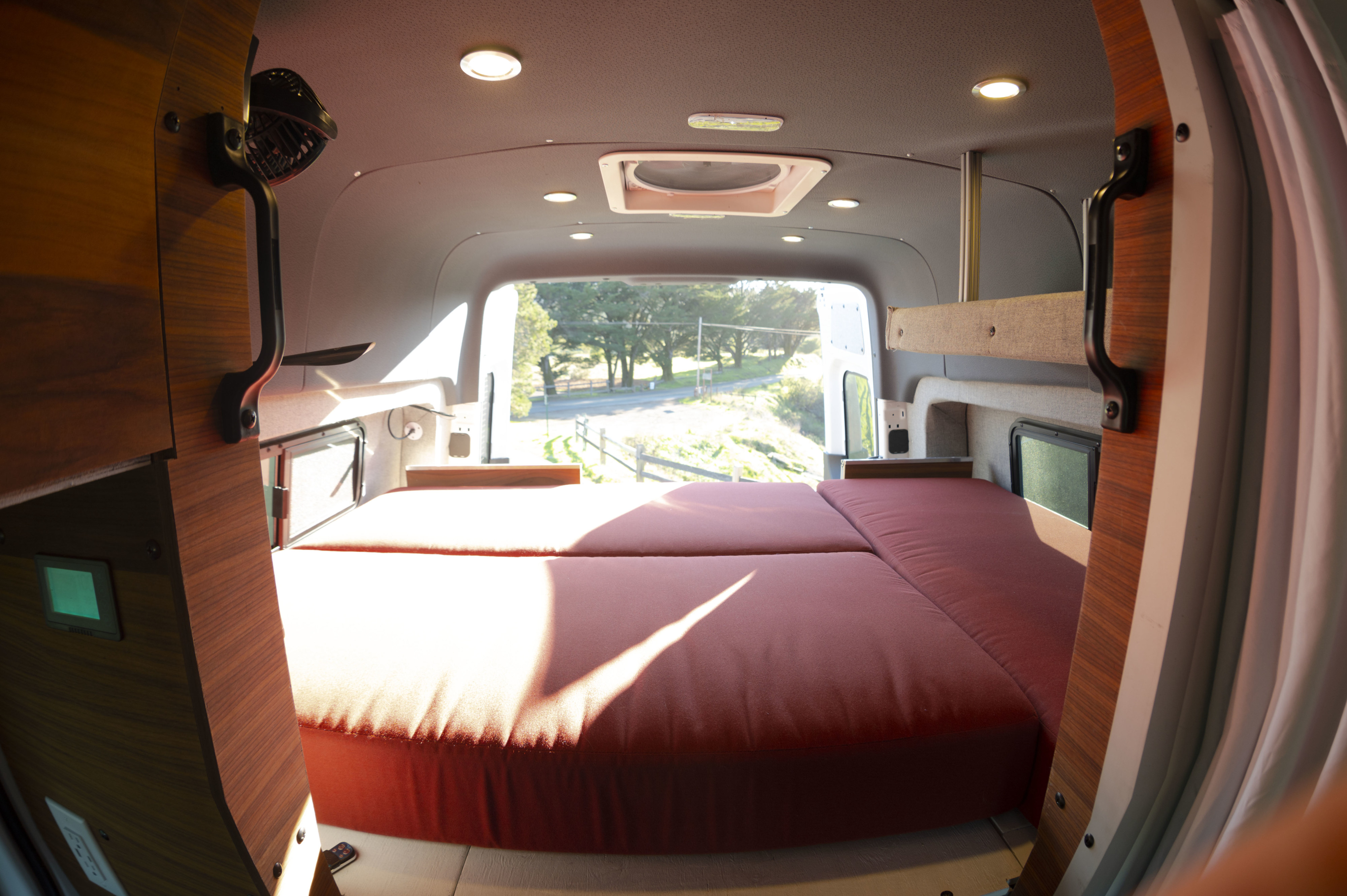 Cozy RV rental interior with a large red bed, wooden details, and a scenic outdoor view through the rear doors.. Mercedes-Benz Sprinter 2019