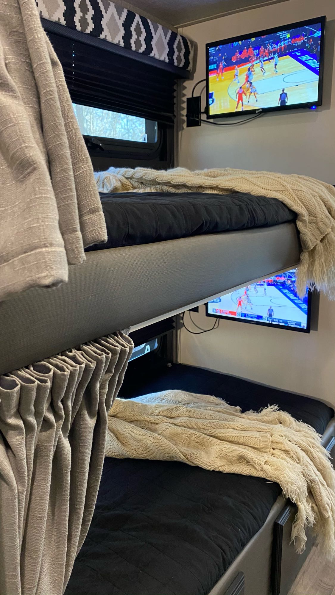 Inside a cozy RV rental: bunk beds with black bedding, cozy throws, and TVs for each bunk showing basketball.. Entegra Coach Odyssey 2022