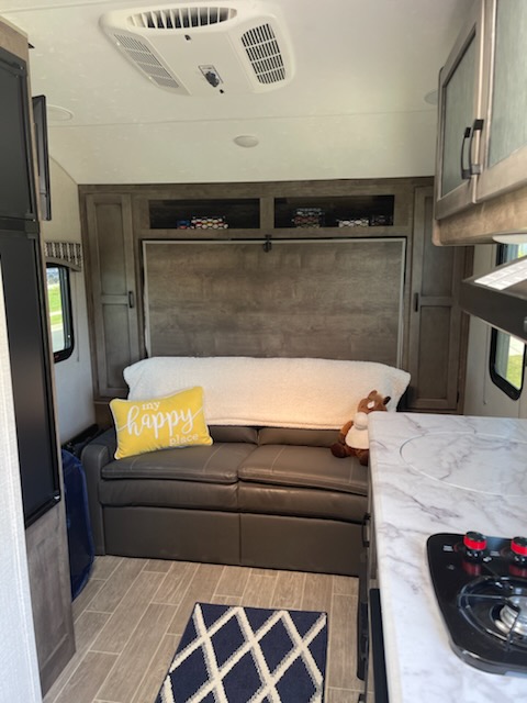 Inside this inviting rv rental, a compact living space features a dark sofa, yellow 'happy place' pillow, kitchen, & ceiling AC.. Sonic Lite 2021