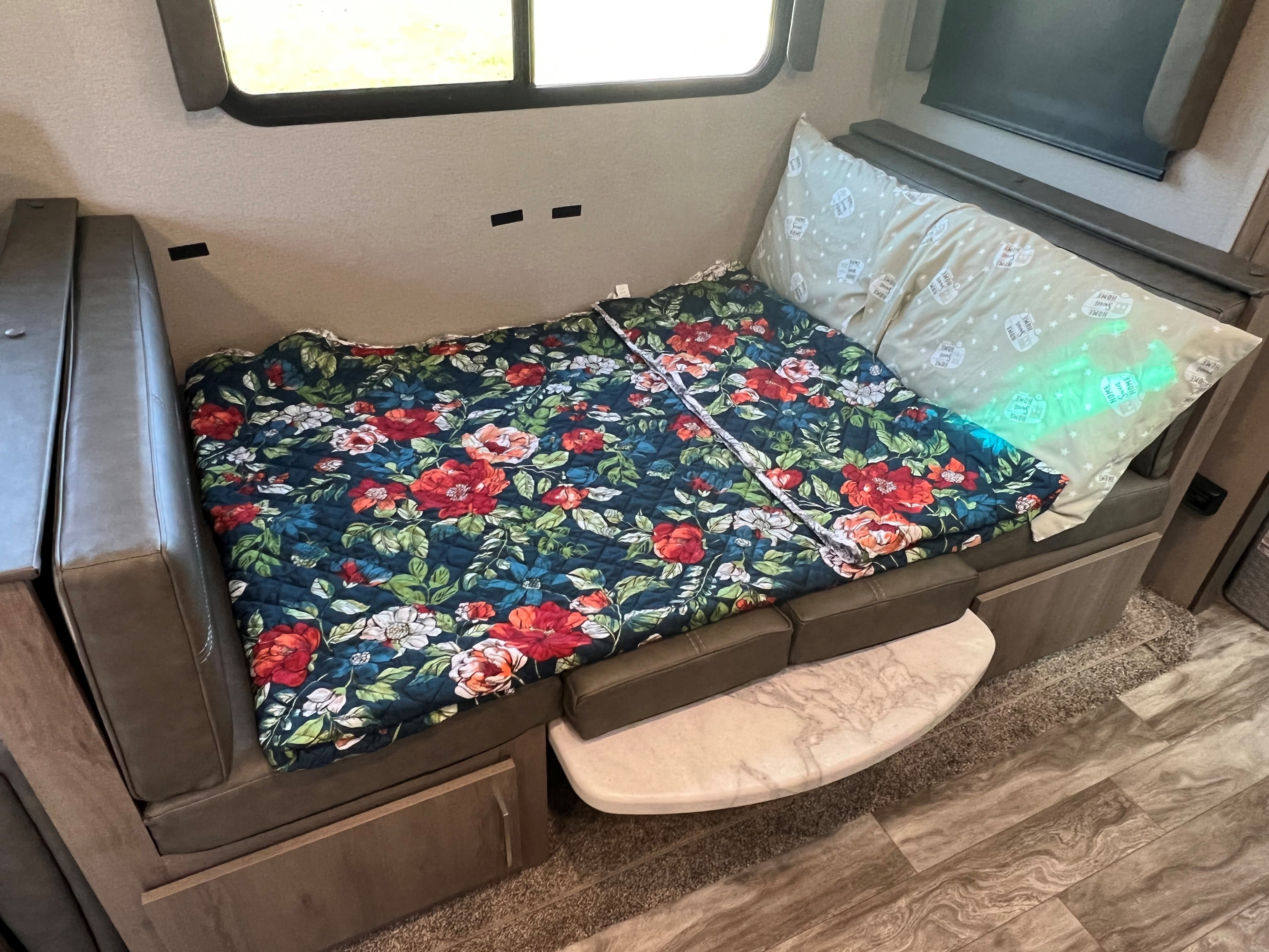 A cozy RV rental dinette transformed into a bed with a vibrant floral blanket and glowing pillows.. Grand Design Imagine 2022