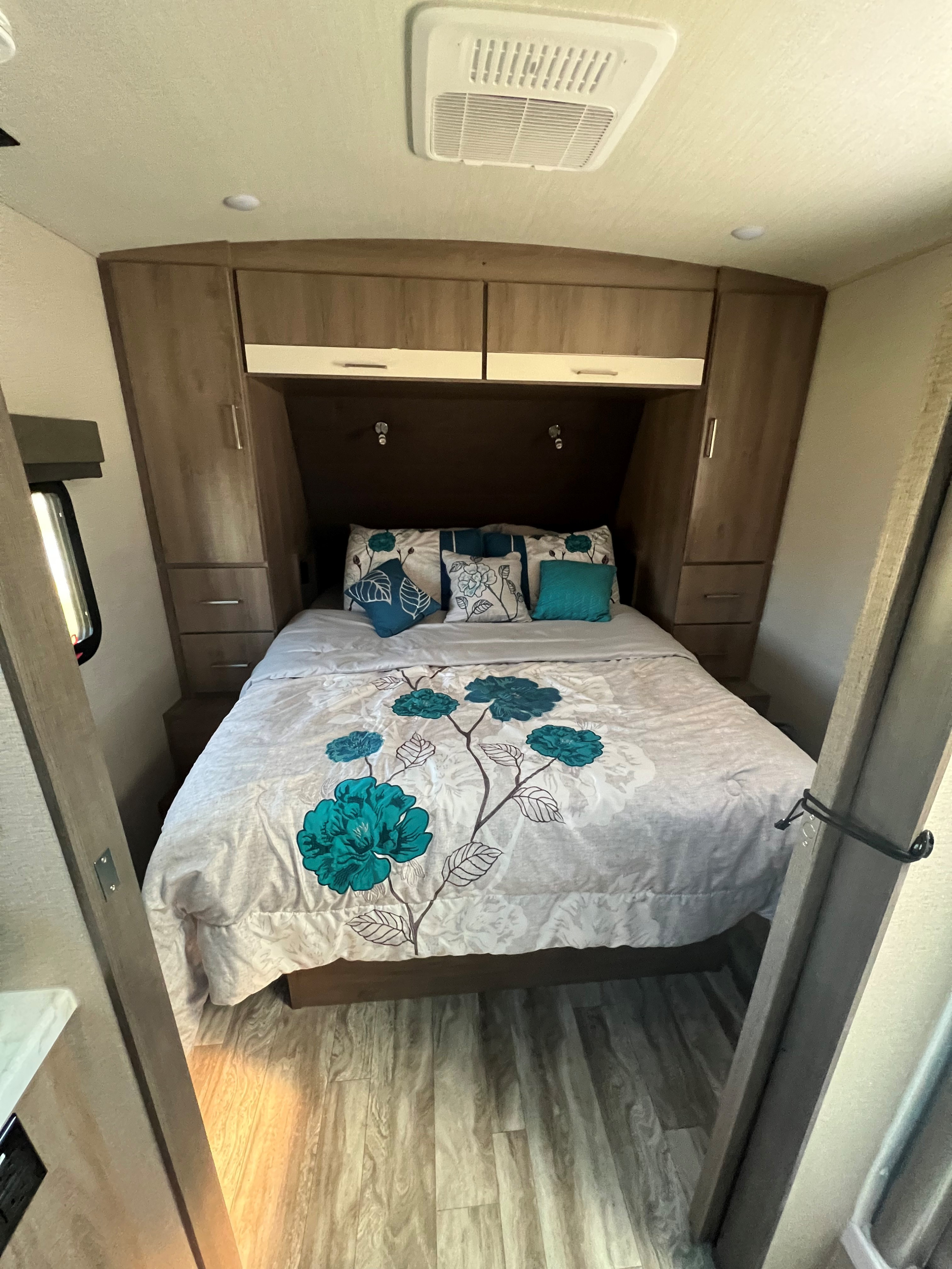 Cozy RV rental bedroom featuring a bed with teal floral bedding, overhead cabinets, and wood flooring for comfortable stays.. Grand Design Imagine 2022