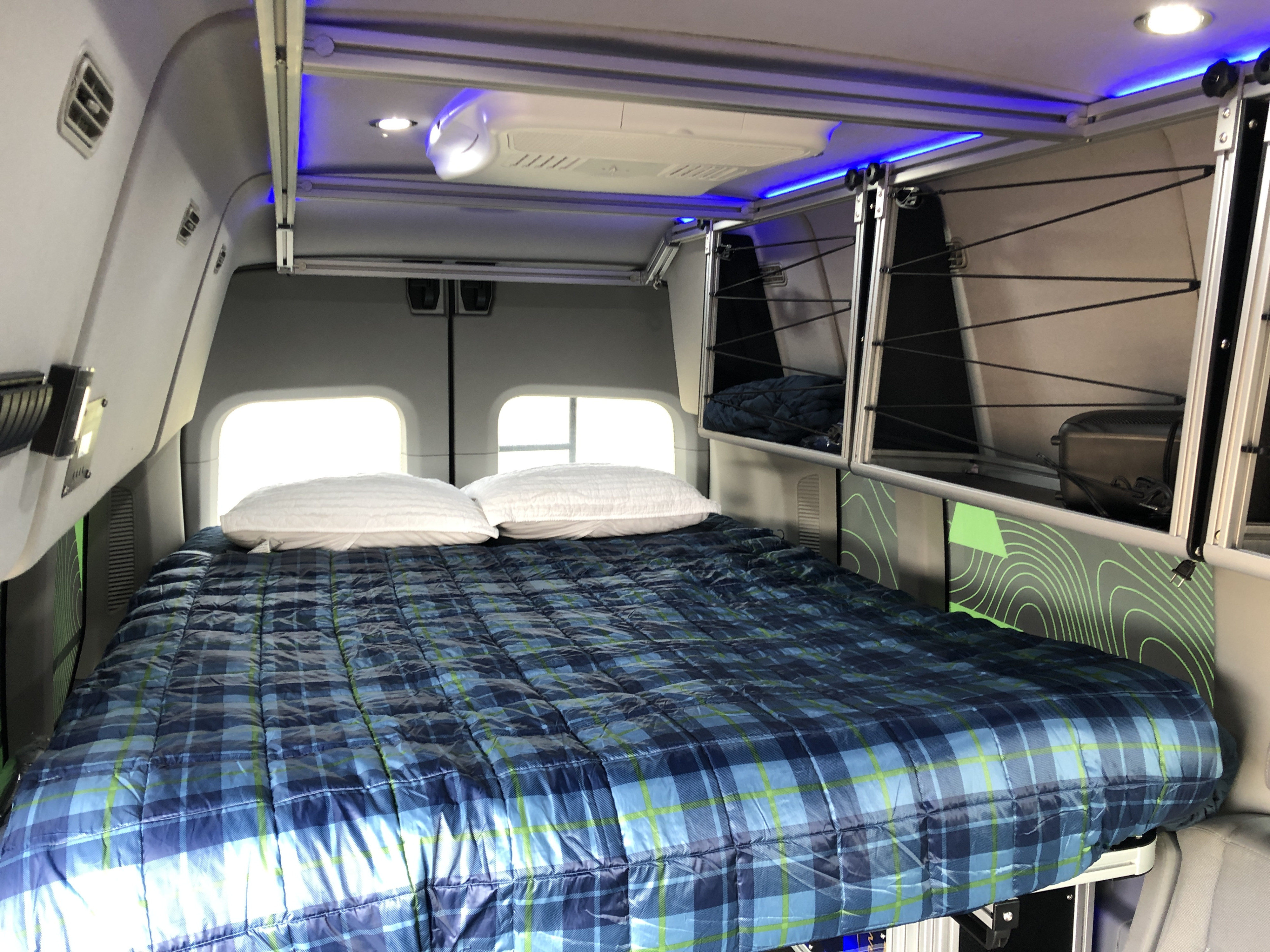 Inside a modern **RV rental**: a plush bed with blue plaid comforter, ample overhead storage, and cool blue ambient lights.. Ford Transit Custom 2017