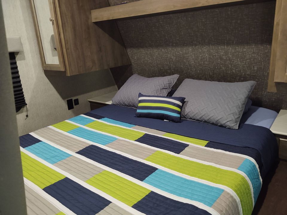 Cozy RV rental bedroom featuring a bed with a vibrant geometric quilt in blue, green, and grey, overhead cabinets.. Heartland RVs Pioneer 2018
