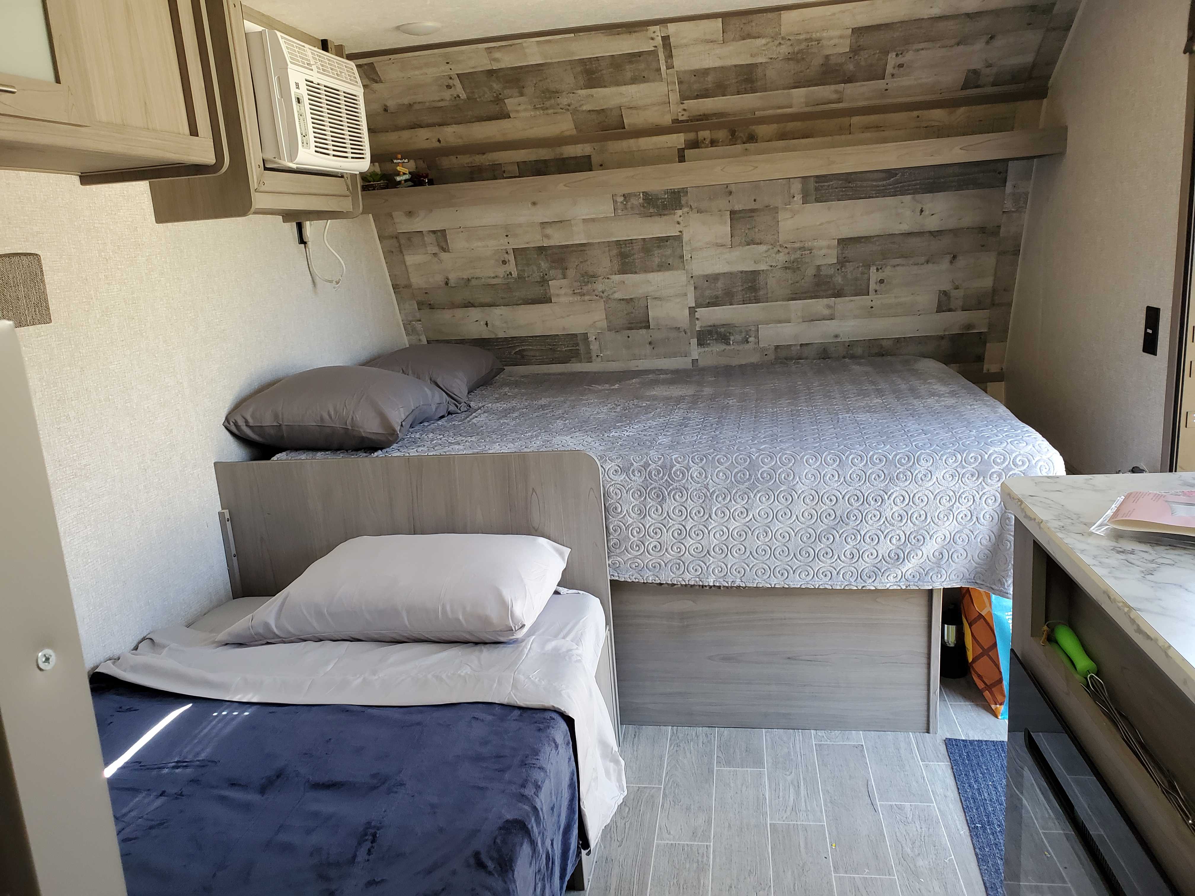 An rv rental bedroom: a large bed with patterned grey bedding, a smaller lower bed, and a wall AC unit. Wood paneling.. Dutchman Colmen 2022