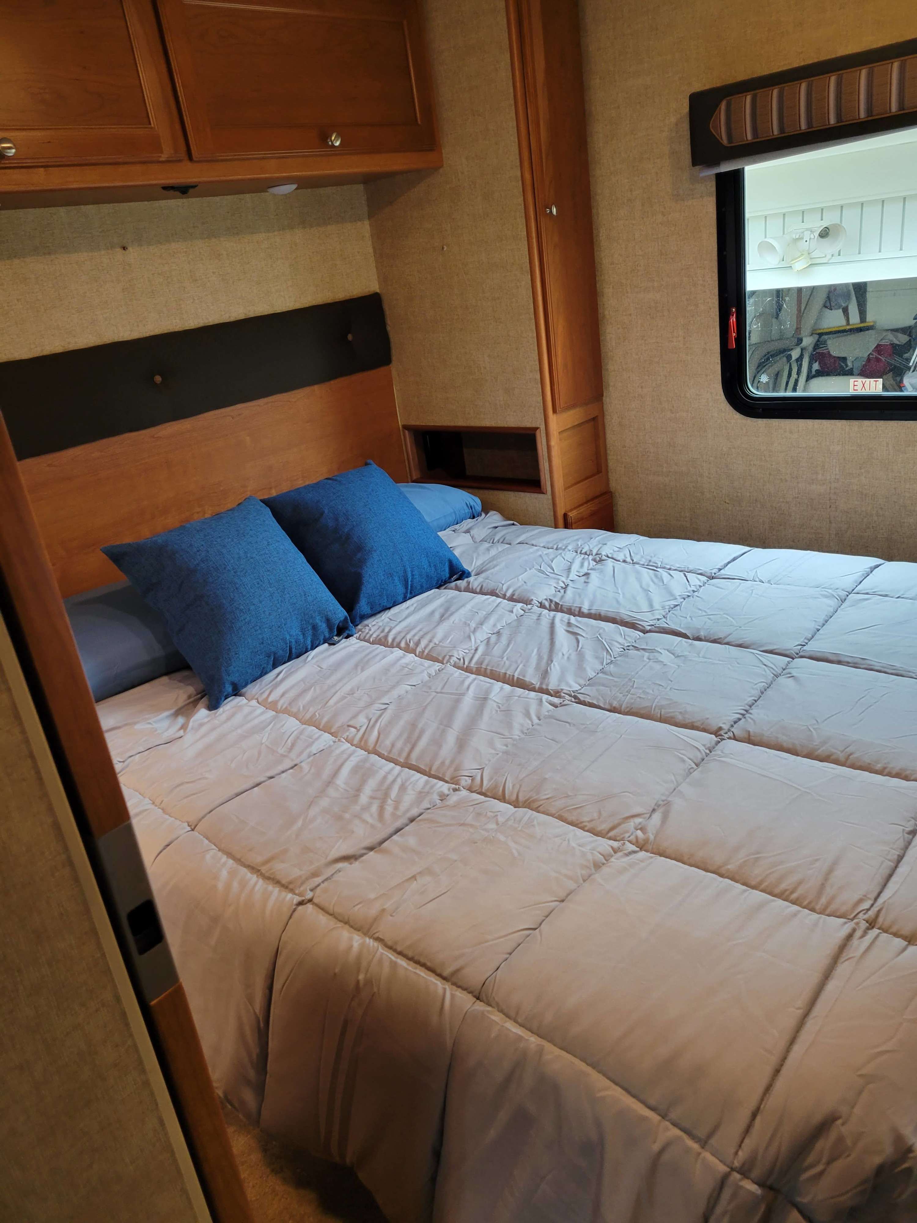 A cozy RV rental bedroom features a queen bed with light grey bedding and blue pillows, plus wooden cabinets and a window.. Winnebago Vista 2018