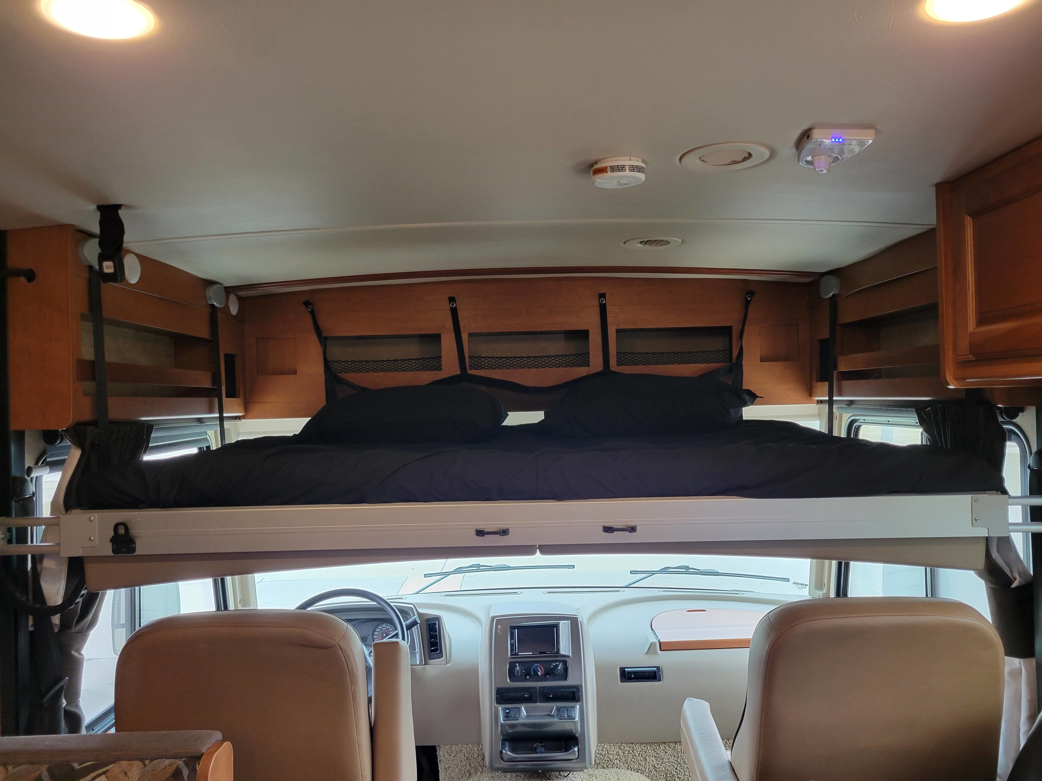 View inside an RV rental: a cozy overhead bunk bed with black bedding sits above the driver's cockpit and dashboard.. Winnebago Vista 2018