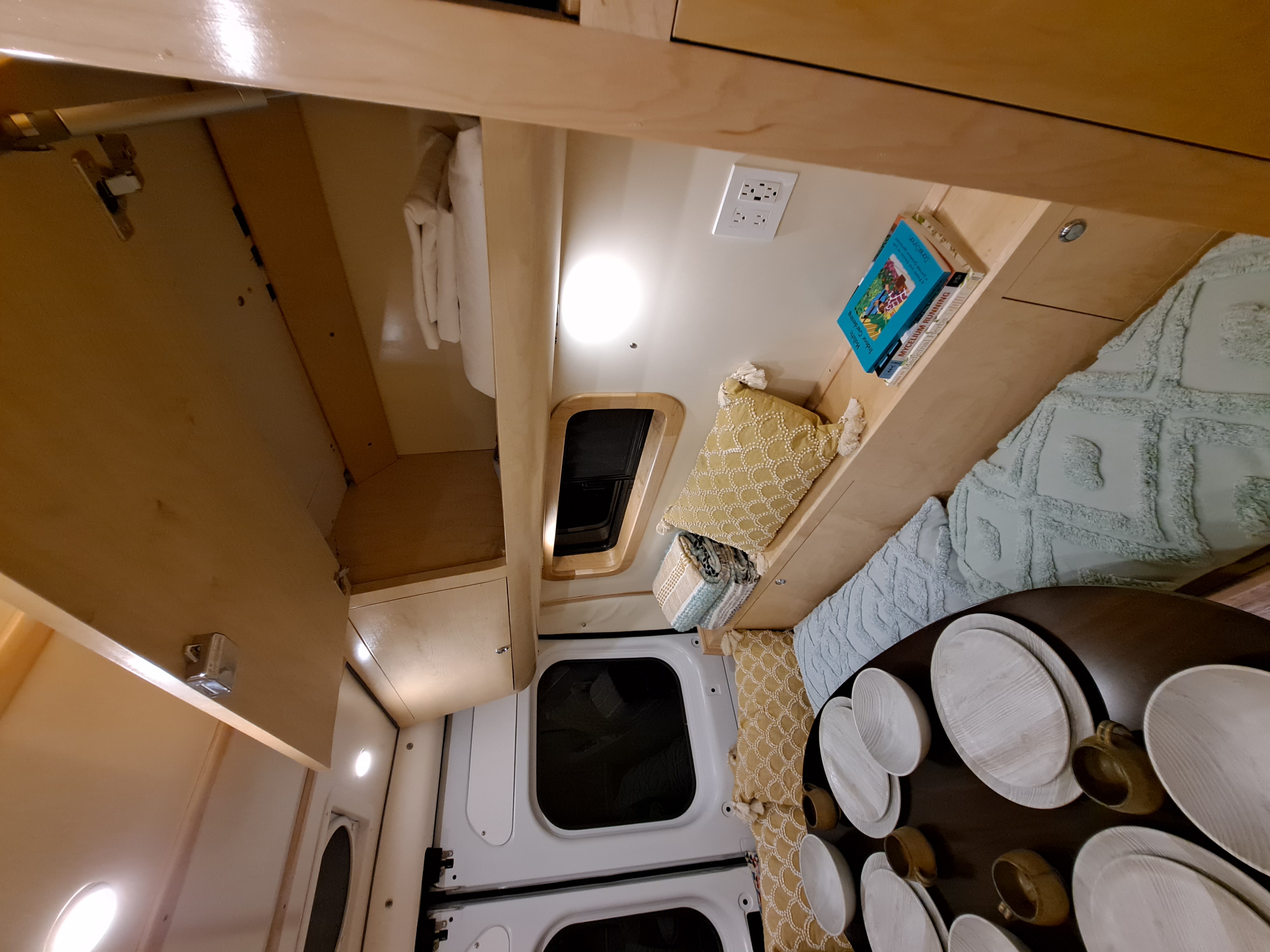 Interior of a modern RV rental, featuring light wooden built-in shelving, a bed with pillows, and a table set with dishes.. Ram ProMaster 3500 2021