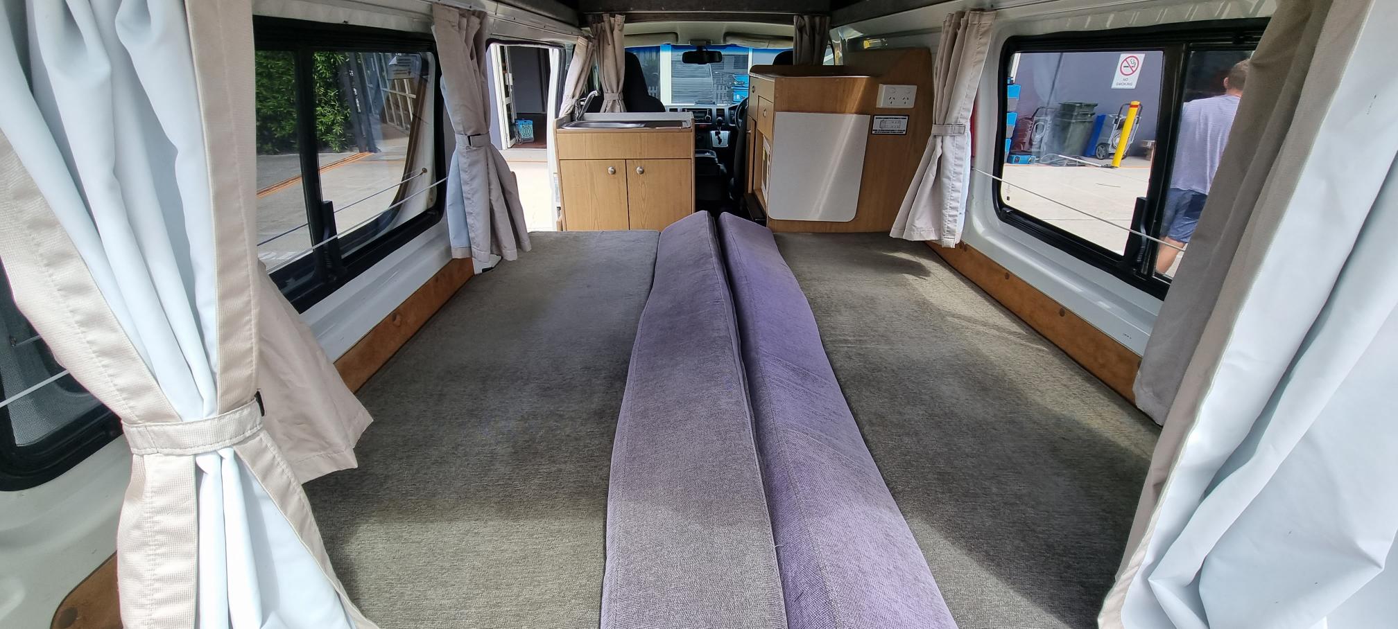 Inside an RV rental: a long, grey carpeted floor, purple cushions forming a bed, small kitchen, and windows with curtains.. Toyota in to the sunset 2014