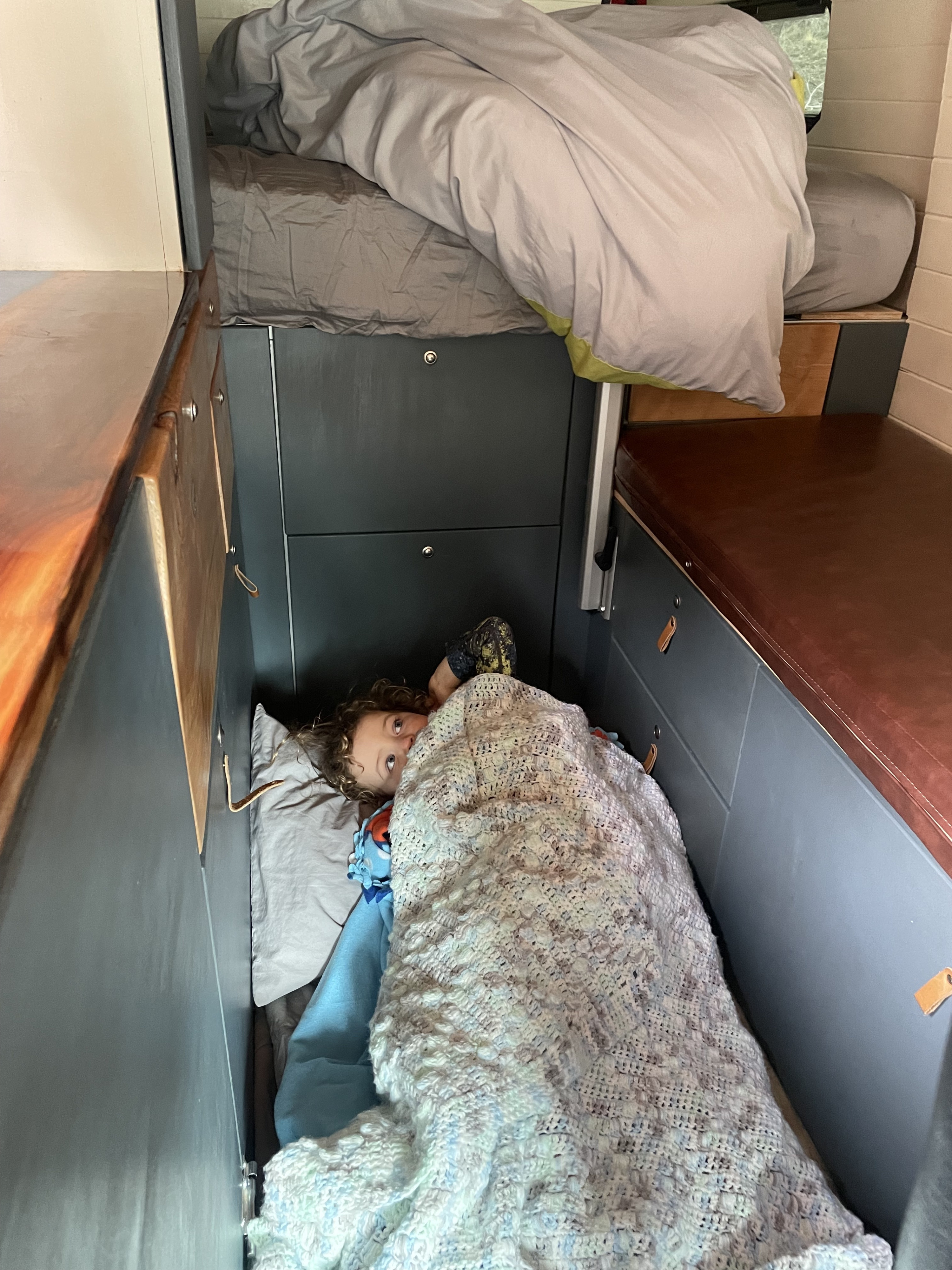 Cozy rv rental bunk: A child peeks from under a blanket in a compact camper van sleeping area, with an upper bunk visible.. Dodge promaster 3500 2018