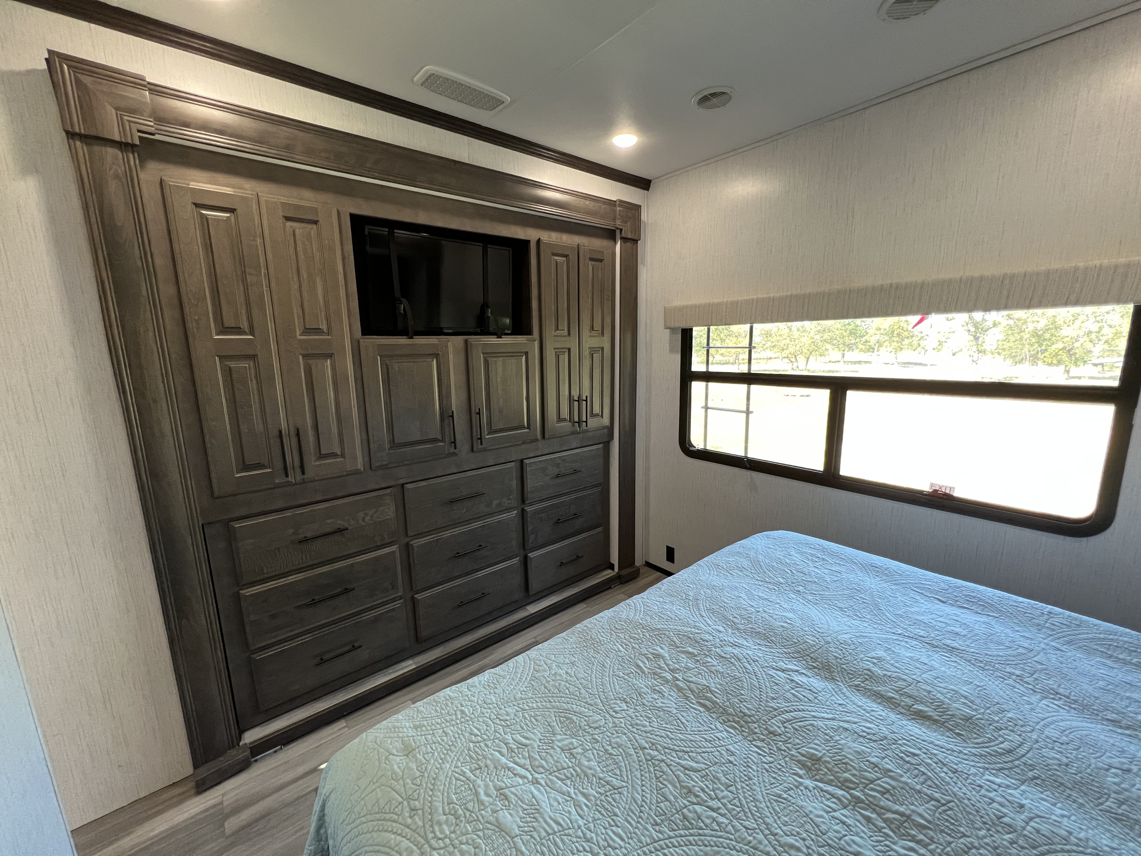 RV rental bedroom interior featuring a large built-in wooden wardrobe with TV, drawers, and a comfortable bed by the window.. Heartland Bighorn 2021
