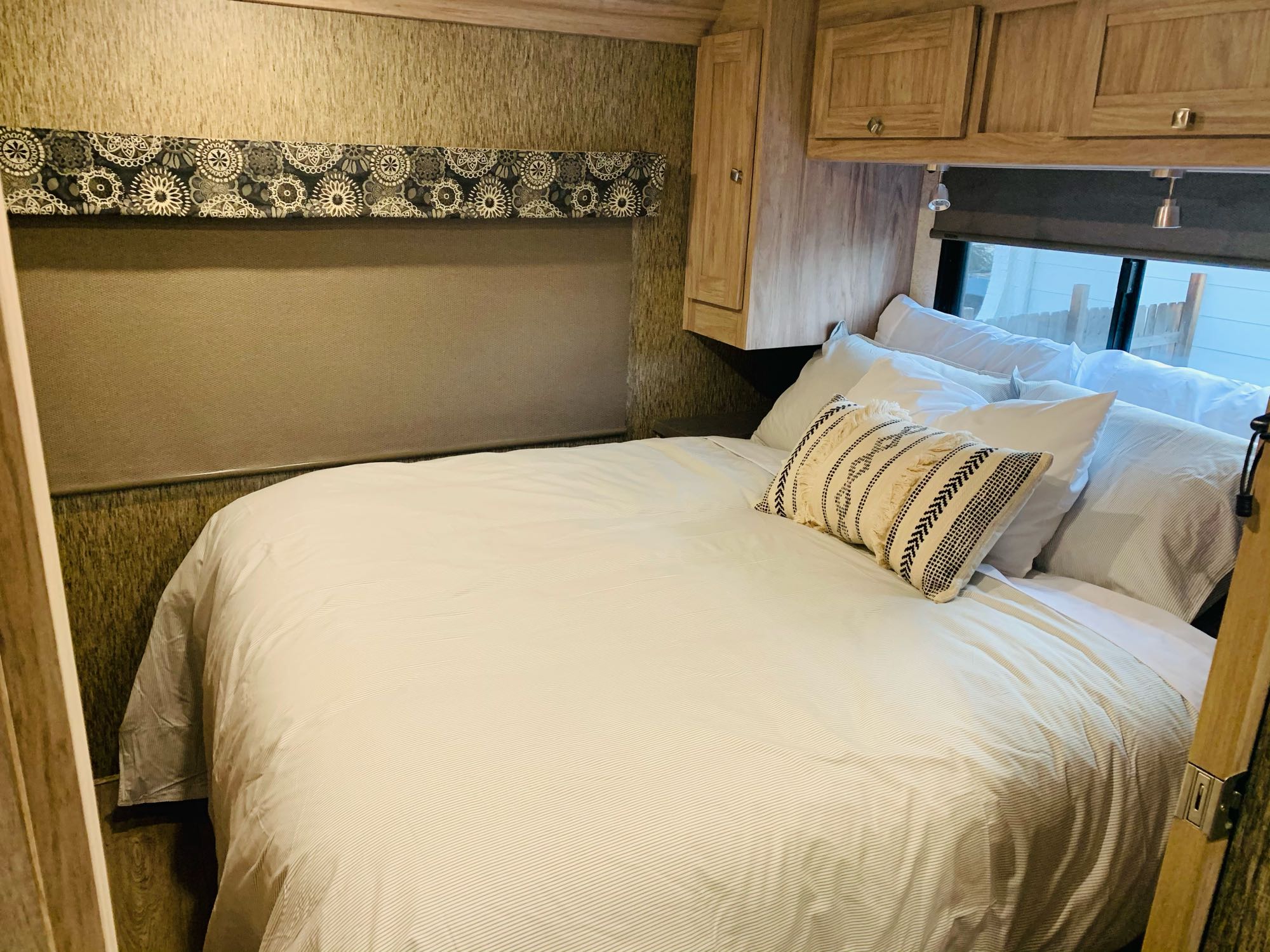 Inside an RV rental: cozy bedroom with striped bedding, multiple pillows, overhead cabinets, and window blind.. Forest River Sunseeker 2017
