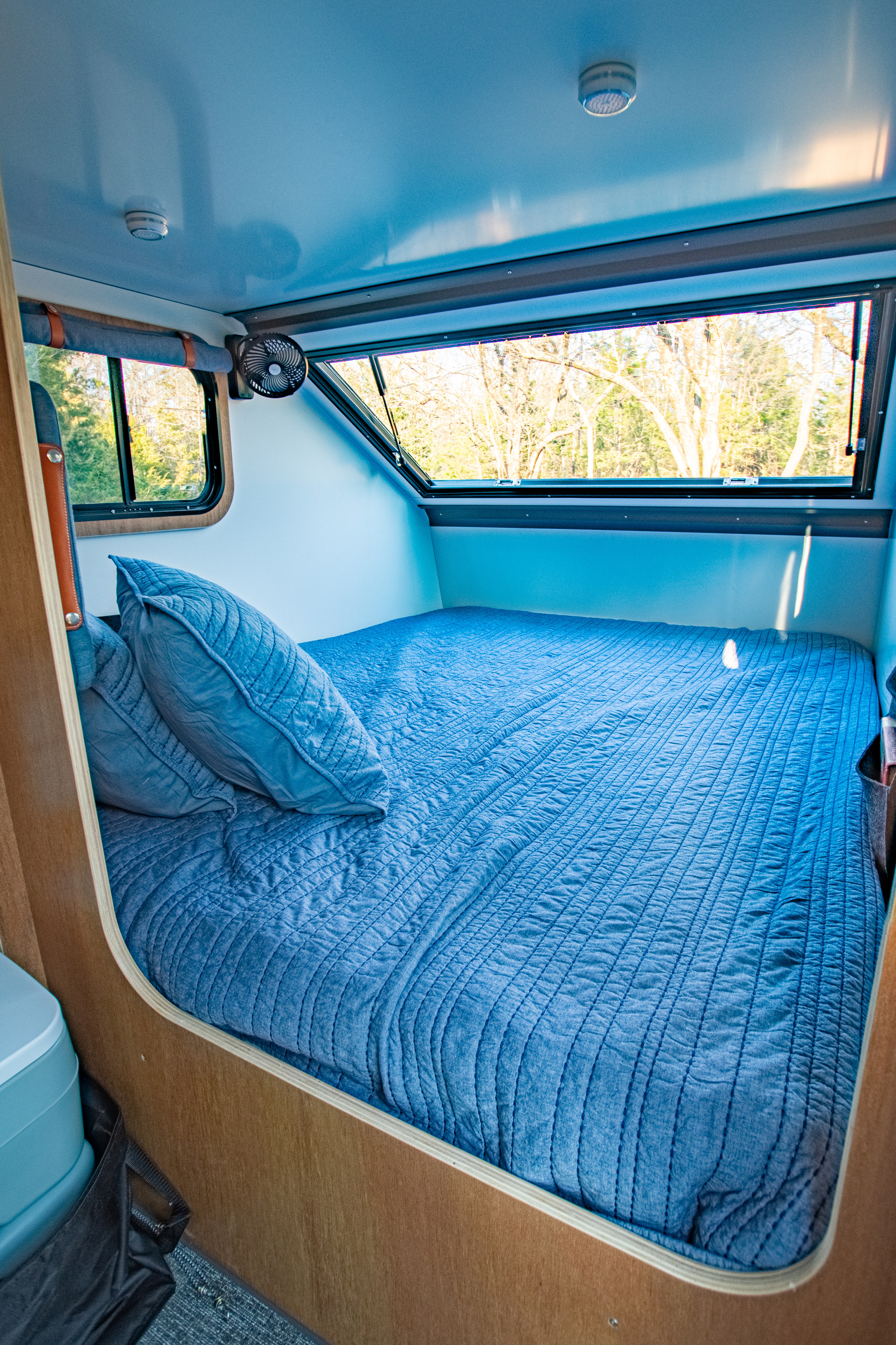 An inviting blue bed inside a compact RV rental, featuring windows with natural views and a ceiling fan.. Campinawe Sport 2022