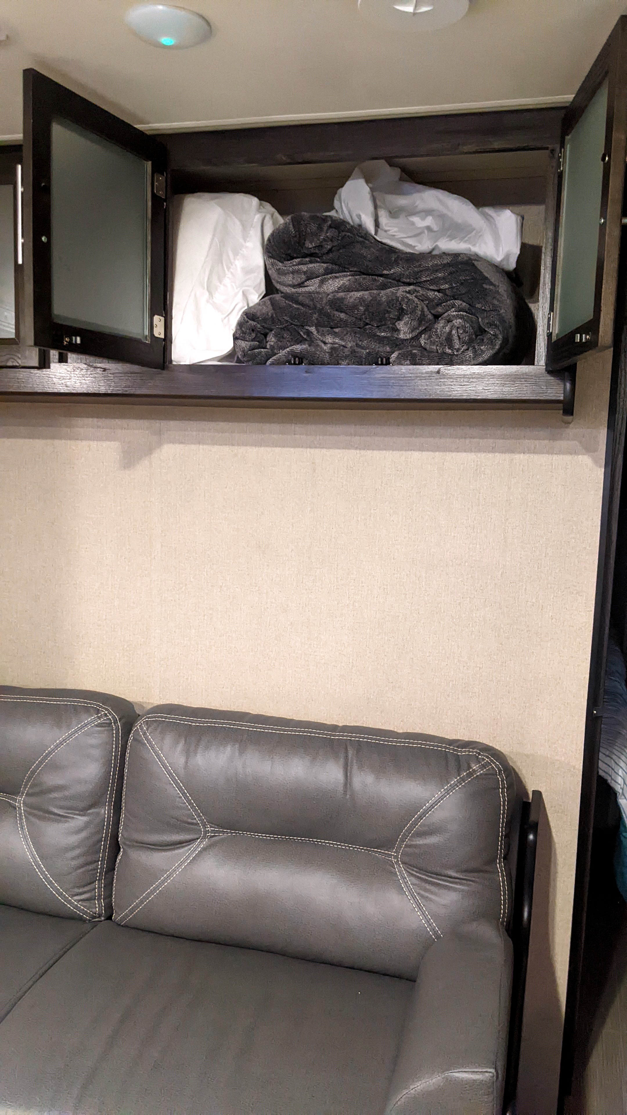 Inside an RV rental: open overhead cabinet with blankets/pillows, ceiling light, and a dark sofa with white stitching.. Dutchmen Kodiak Express 2017