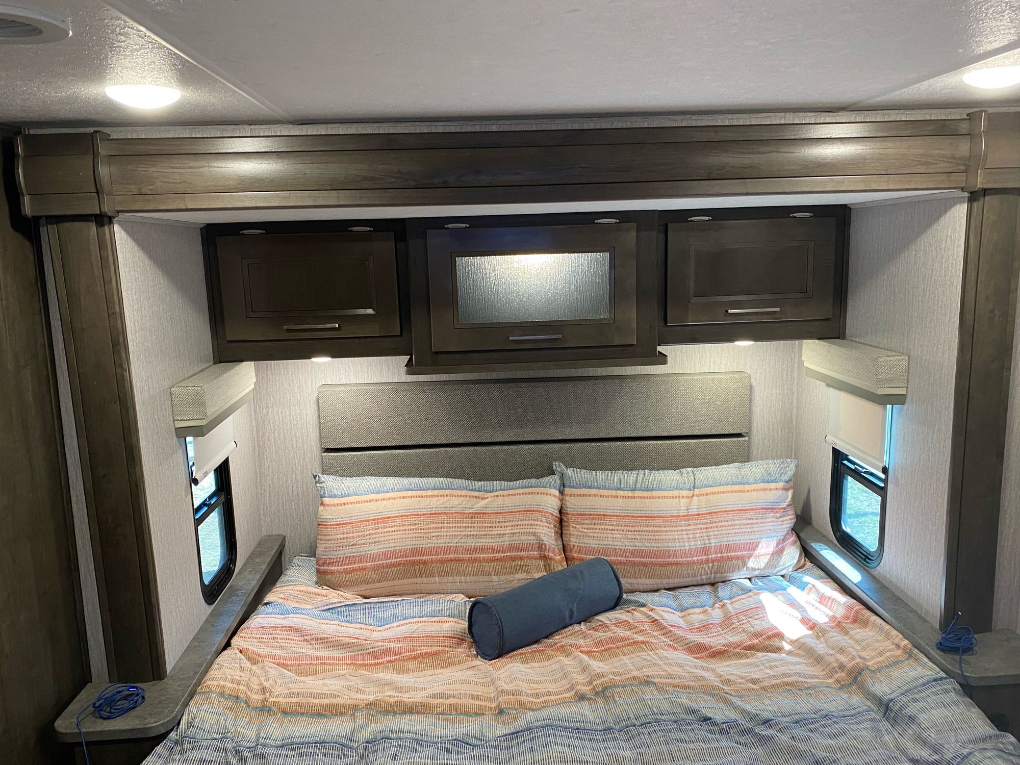 Relax in this cozy RV rental bedroom featuring a striped bed, headboard, overhead cabinets, and side windows.. Forest River Coachmen Mirada 2022