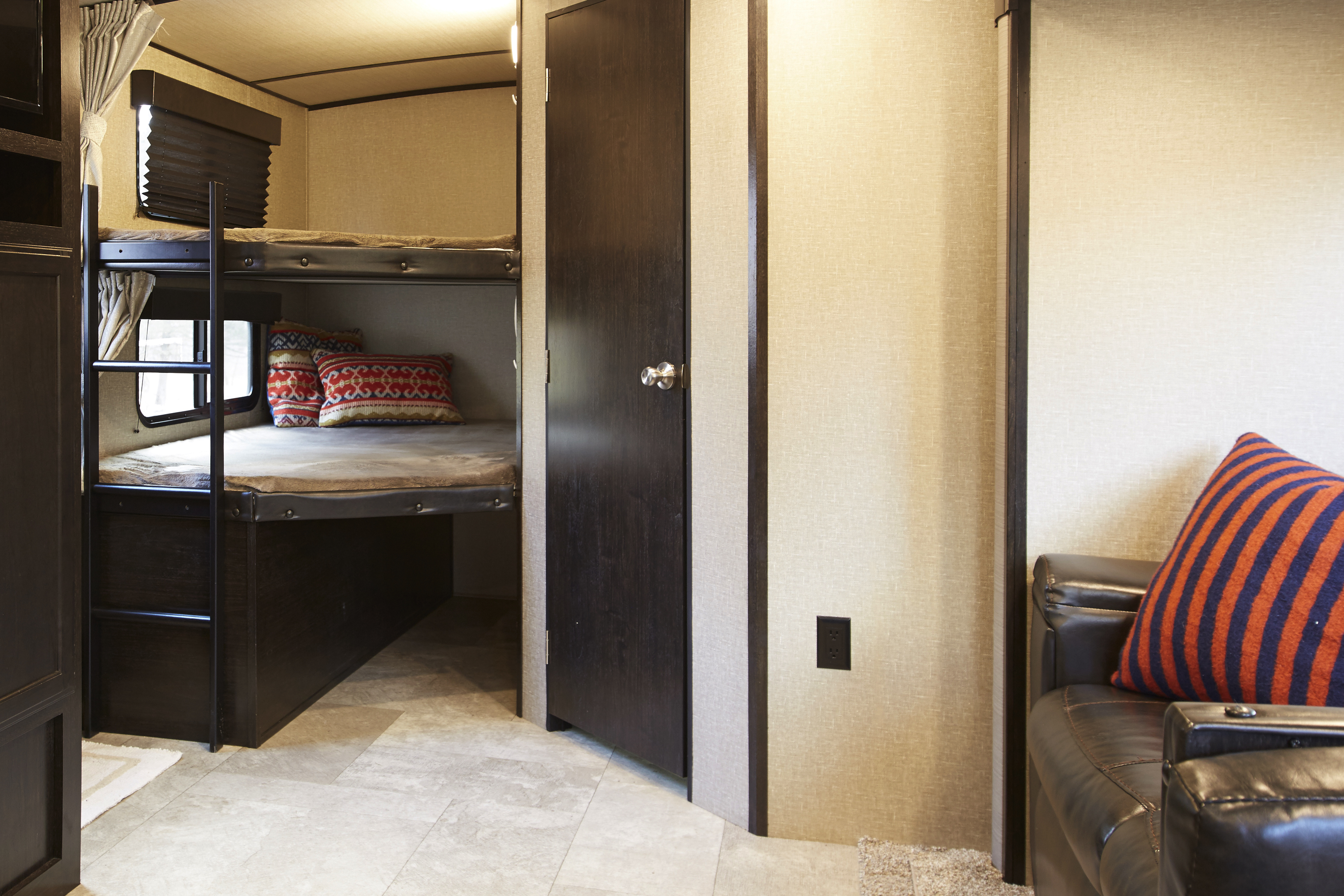 Inside an RV rental, showcasing bunk beds with patterned pillows, a dark wood door, and a comfy brown leather couch.. Grand Design Imagine 2019