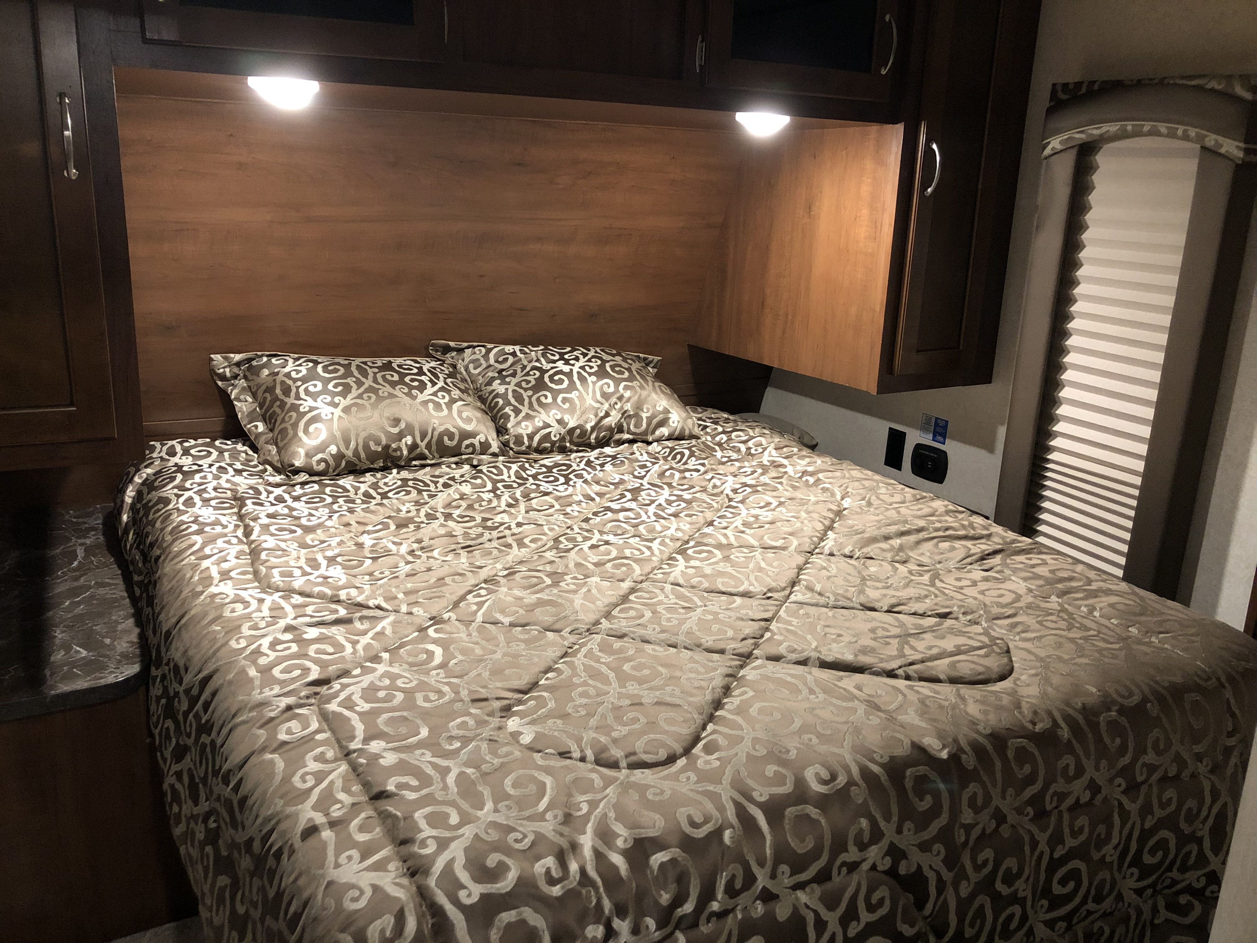RV rental bedroom with patterned bed, reading lights, overhead cabinets, and a window with blinds.. Jayco Jay Flight 2017