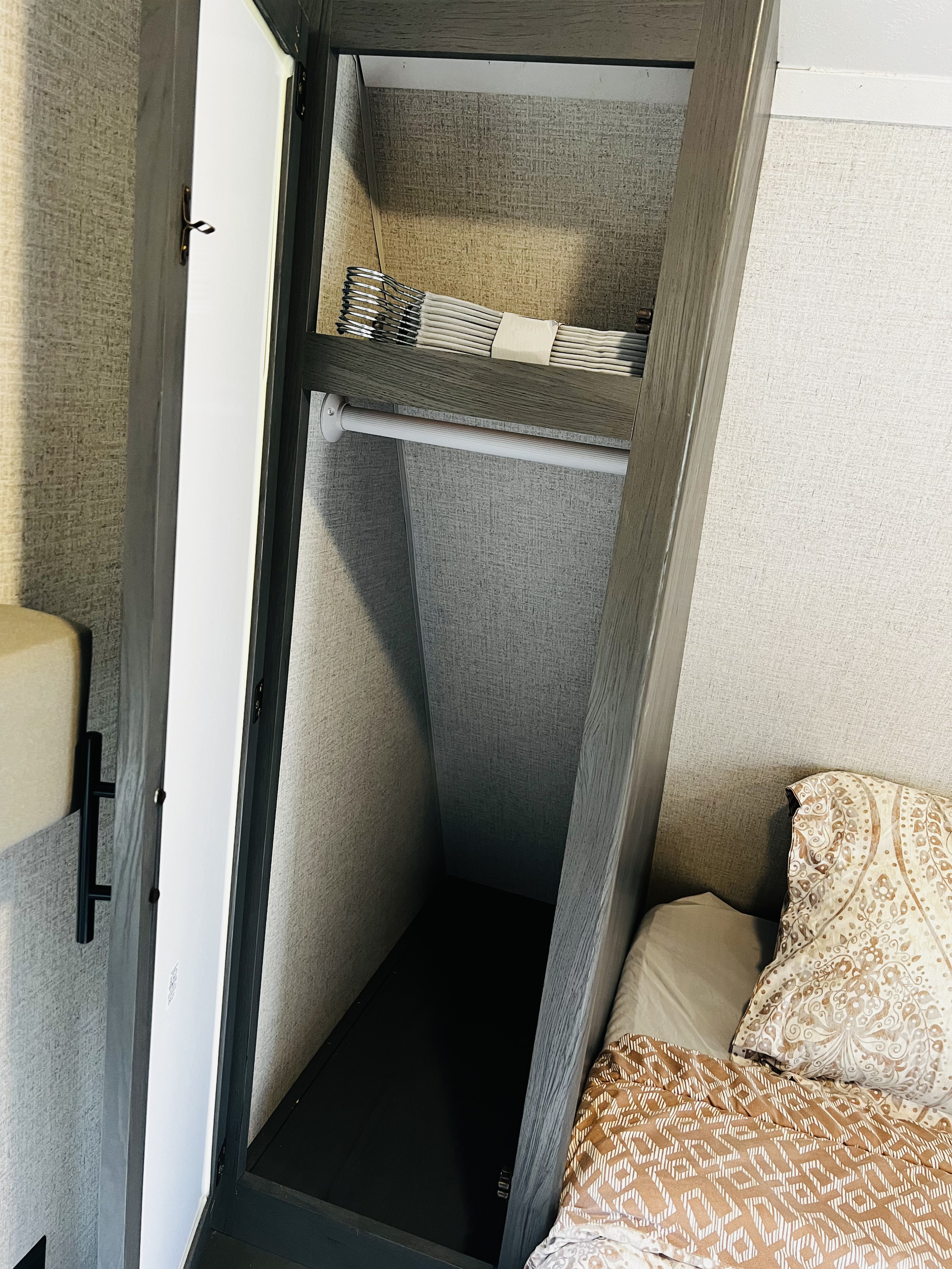 Inside an rv rental: a compact closet with hanging rod and shelf holding neatly folded items, next to the bed.. Dutchmen Aspen Trail LE 2023
