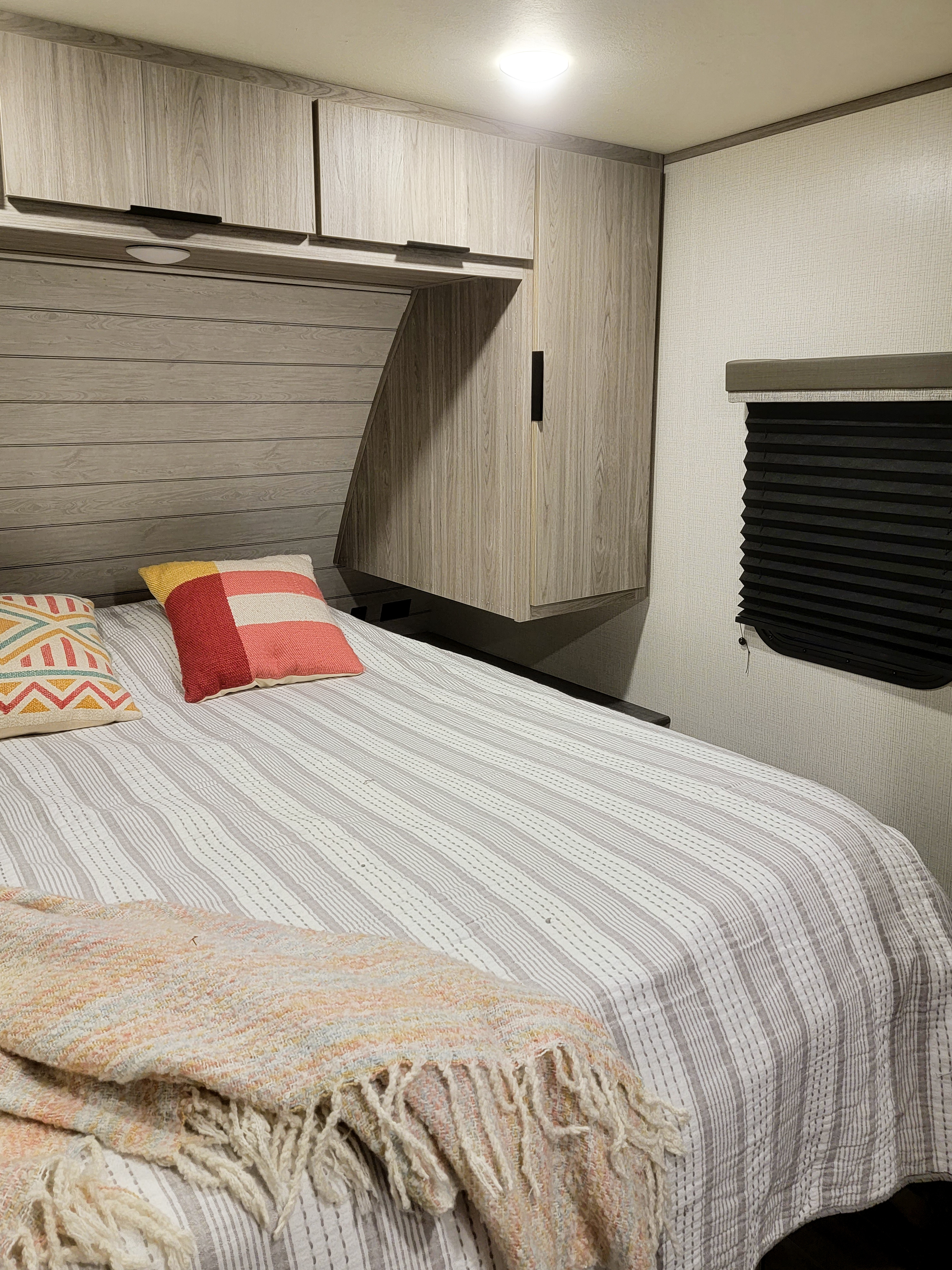 Modern RV rental bedroom interior with light wood cabinets, striped bedding, colorful pillows, and a window blind.. Keystone RV Passport SL 2021
