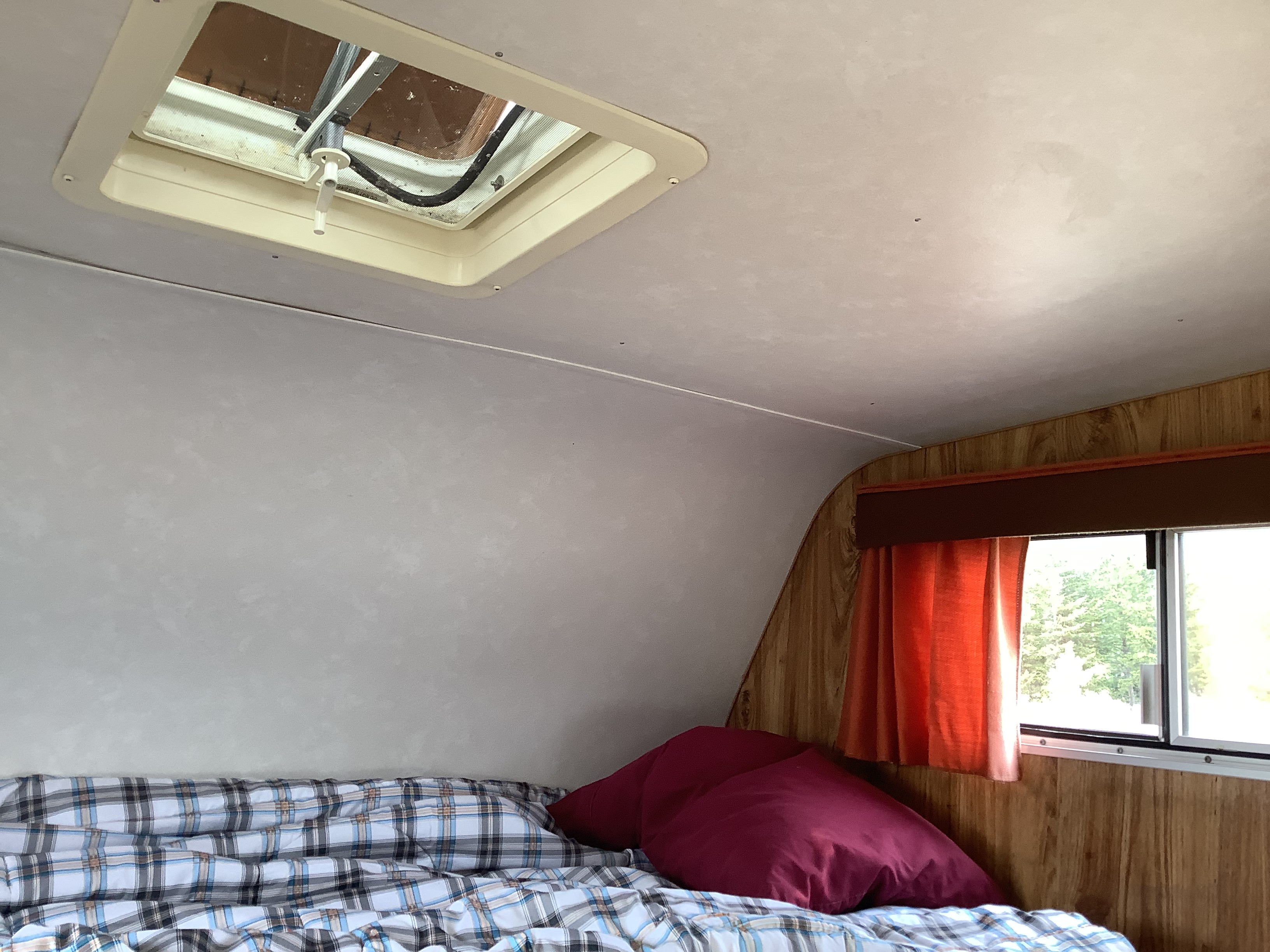 Inside an RV rental: Bed with plaid bedding & red pillows, wood walls, window with orange curtains, and a ceiling vent.. Vanguard Industries MV 1800 1981