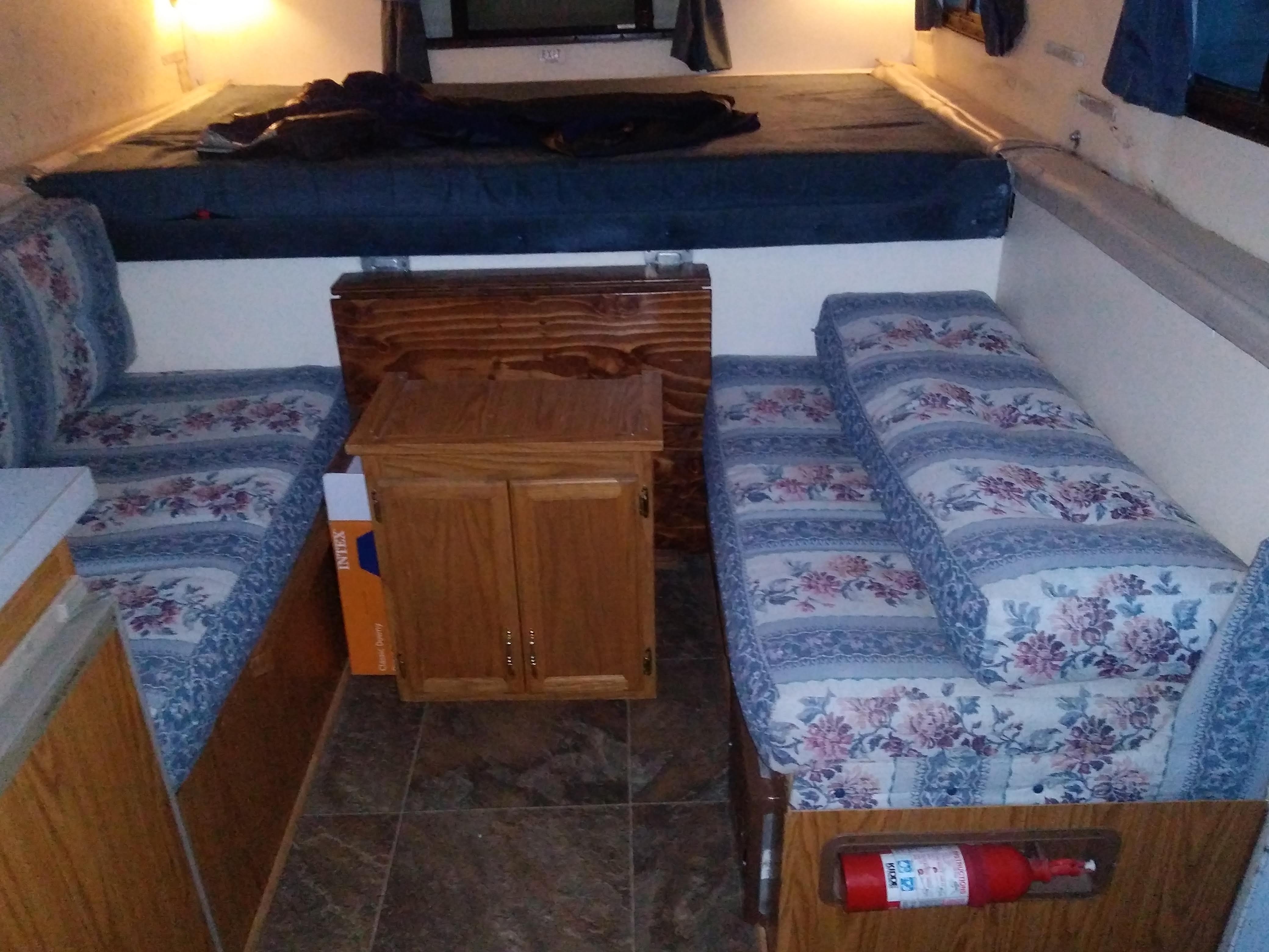 RV rental interior featuring a bed, floral dinette seating, and a wooden cabinet. Fire extinguisher visible for safety.. Trailmanor 2619 1996