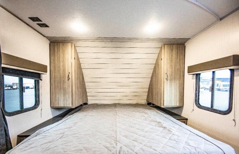 Cozy RV rental bedroom with a large bed centered between angled wood cabinets, windows, and overhead lighting.. Keystone RV Passport SL 2021