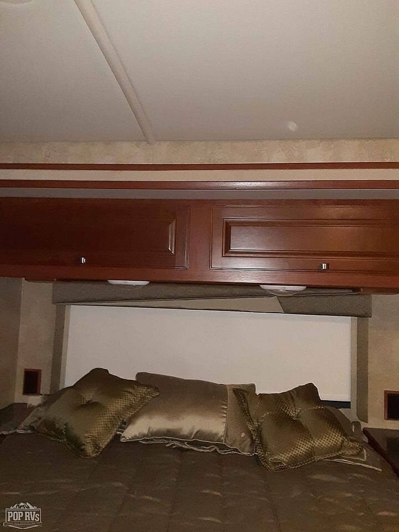View of an RV rental's bedroom, featuring a comfortable bed below functional overhead wooden cabinets for storage.. Winnebago Vista 2014