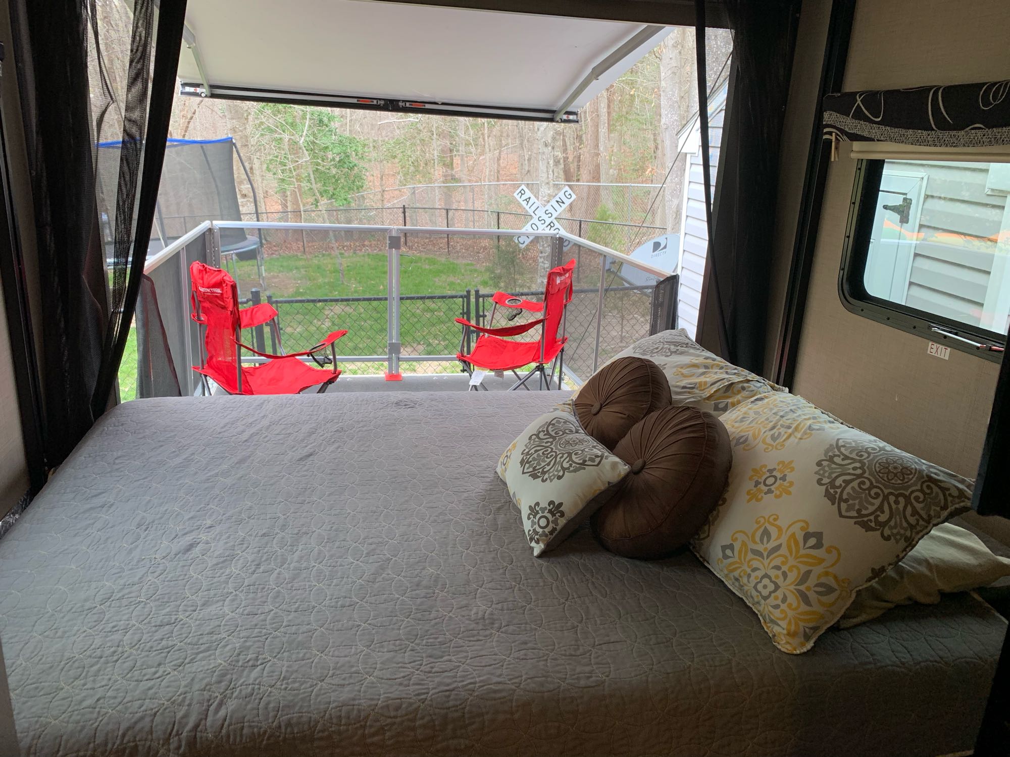 Relaxing RV rental view: comfortable bed facing an outdoor patio with red chairs, a backyard, and a railroad crossing sign.. Thor Outlaw 2018
