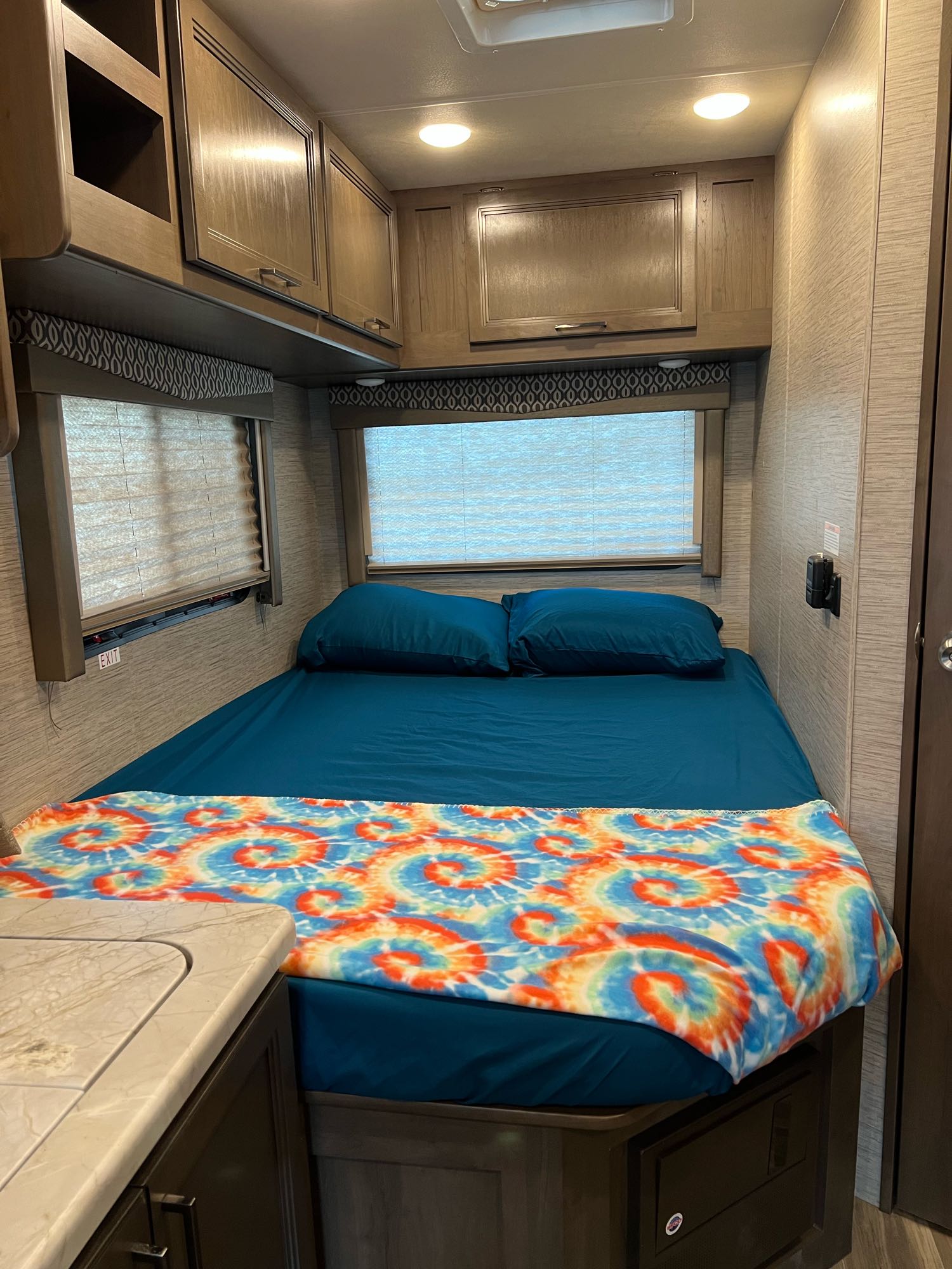 Compact RV rental bedroom with a teal bed, colorful tie-dye blanket, and ample overhead cabinets.. Thor Freedom Elite 2021