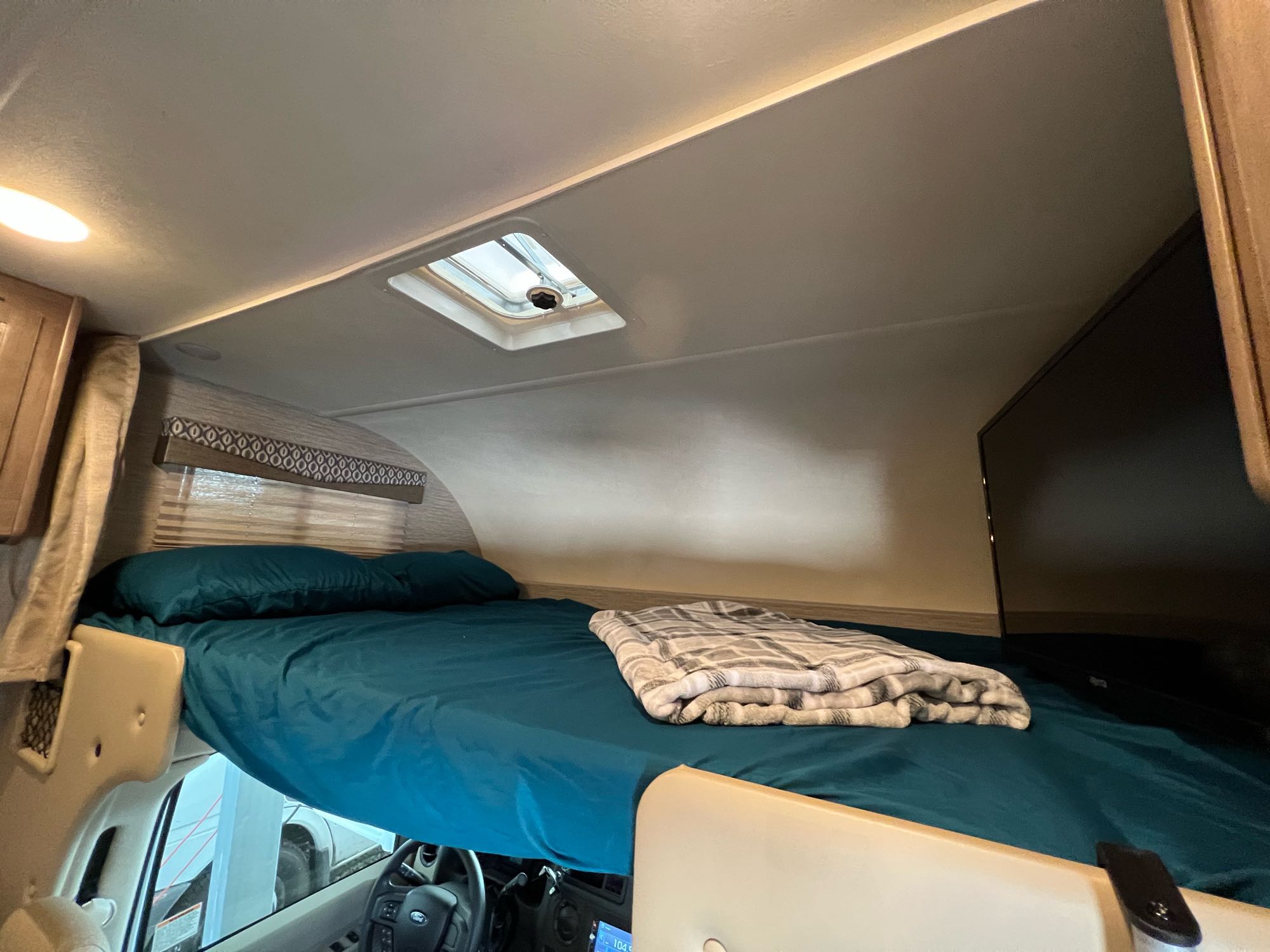 Cozy RV rental cab-over bed features teal sheets, blanket, skylight, and mounted TV, perfect for motorhome travel.. Thor Freedom Elite 2021