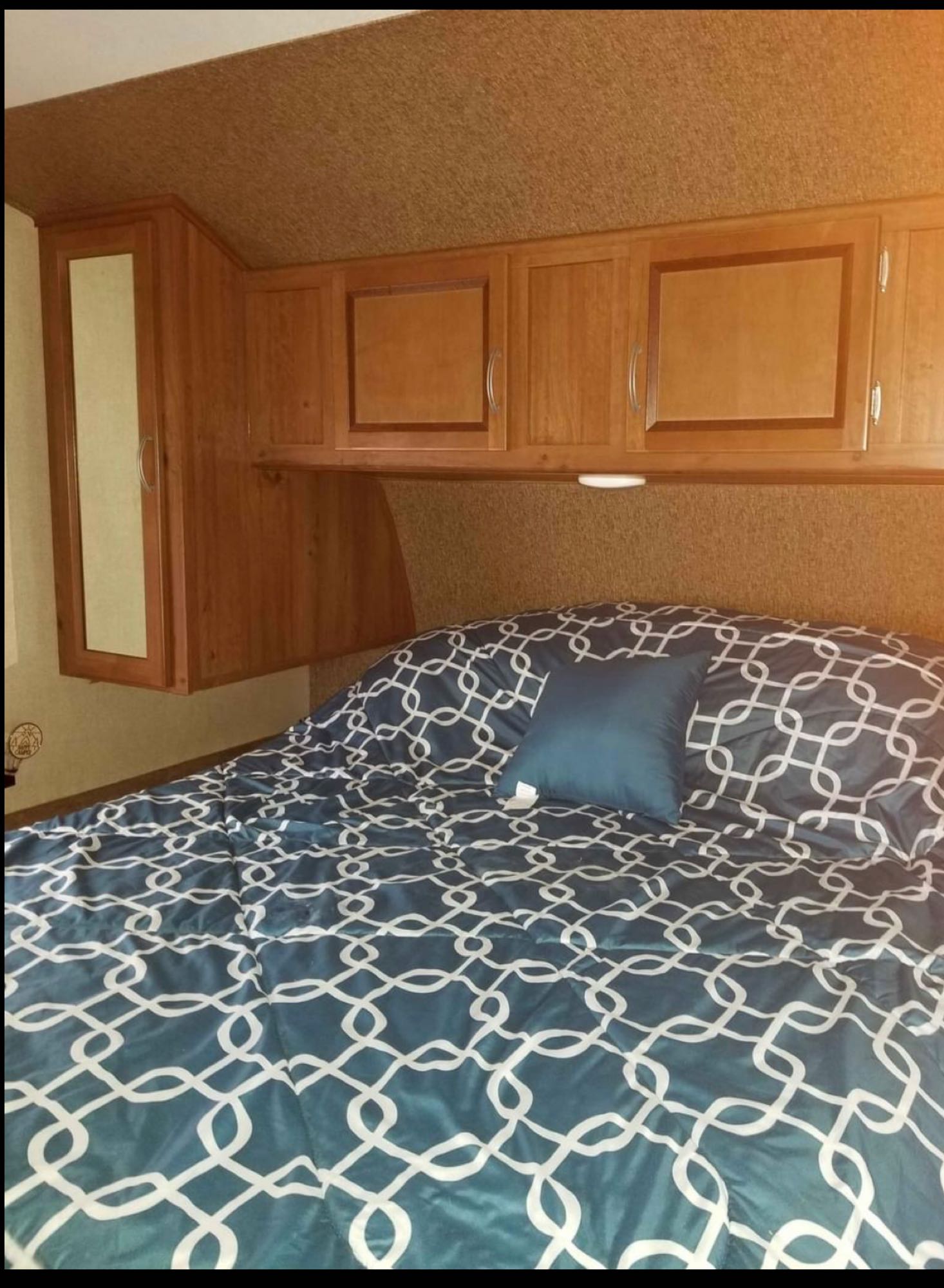 This cozy RV rental bedroom features a bed with blue patterned bedding, a small pillow, and overhead wooden cabinets.. Gulf Stream Innsbruck 2017