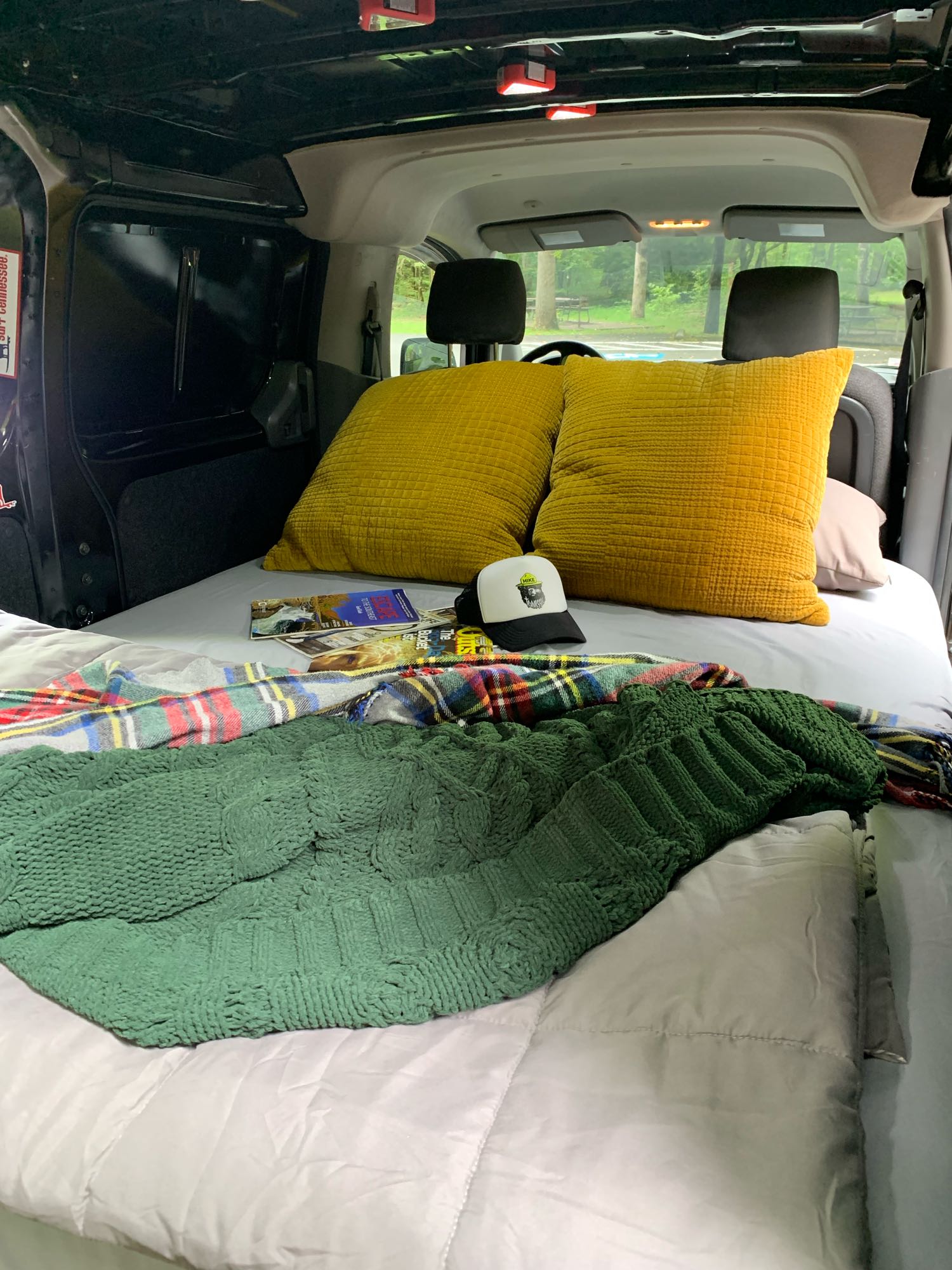 Cozy RV rental bed with yellow pillows, plaid & green blankets, magazines, and a hat, ready for travel.. Nissan Other 2017