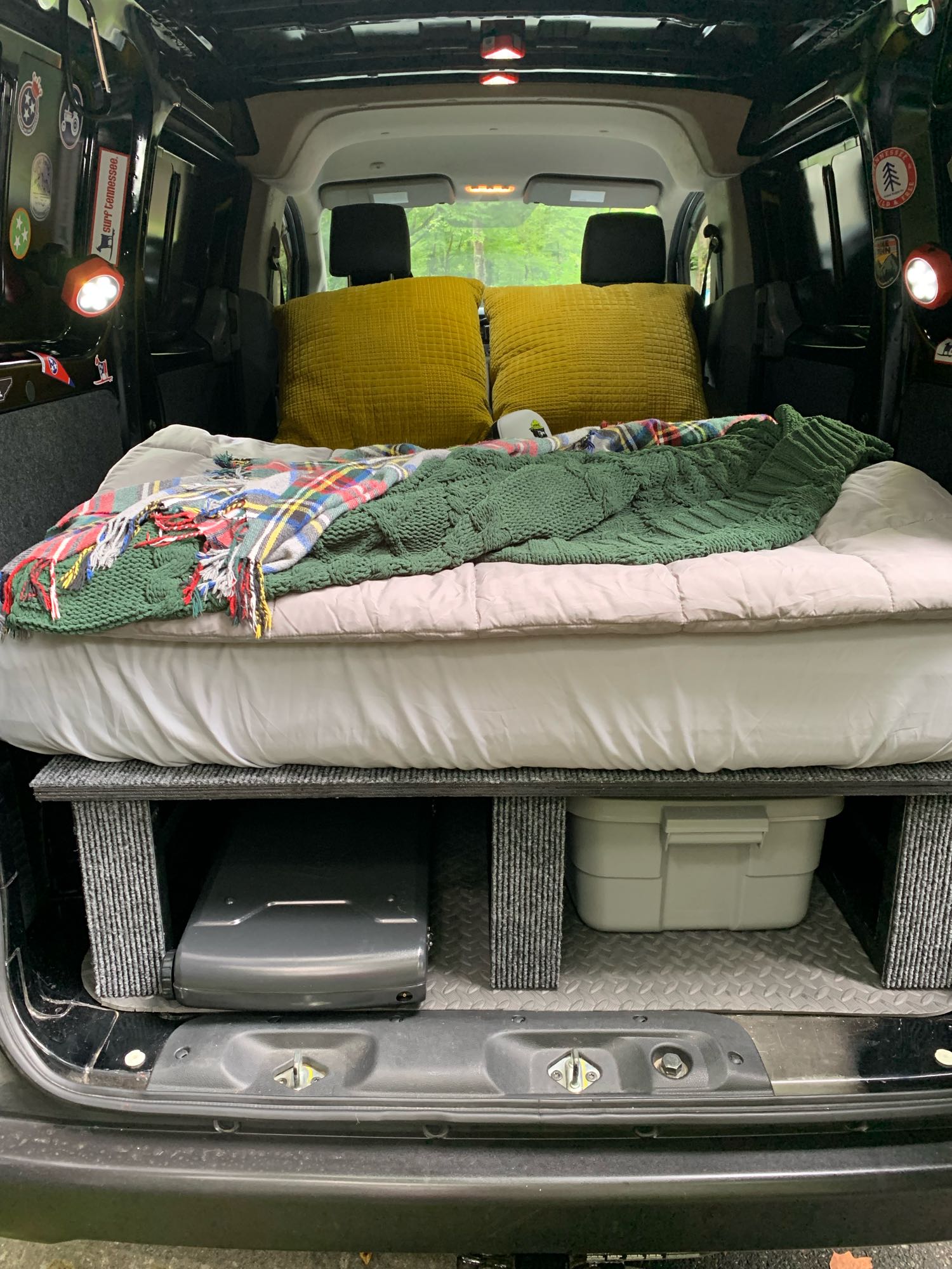 Inside a DIY rv rental, a comfortable bed sits over under-bed storage for gear. Yellow pillows and blankets create a cozy camp setup.. Nissan Other 2017