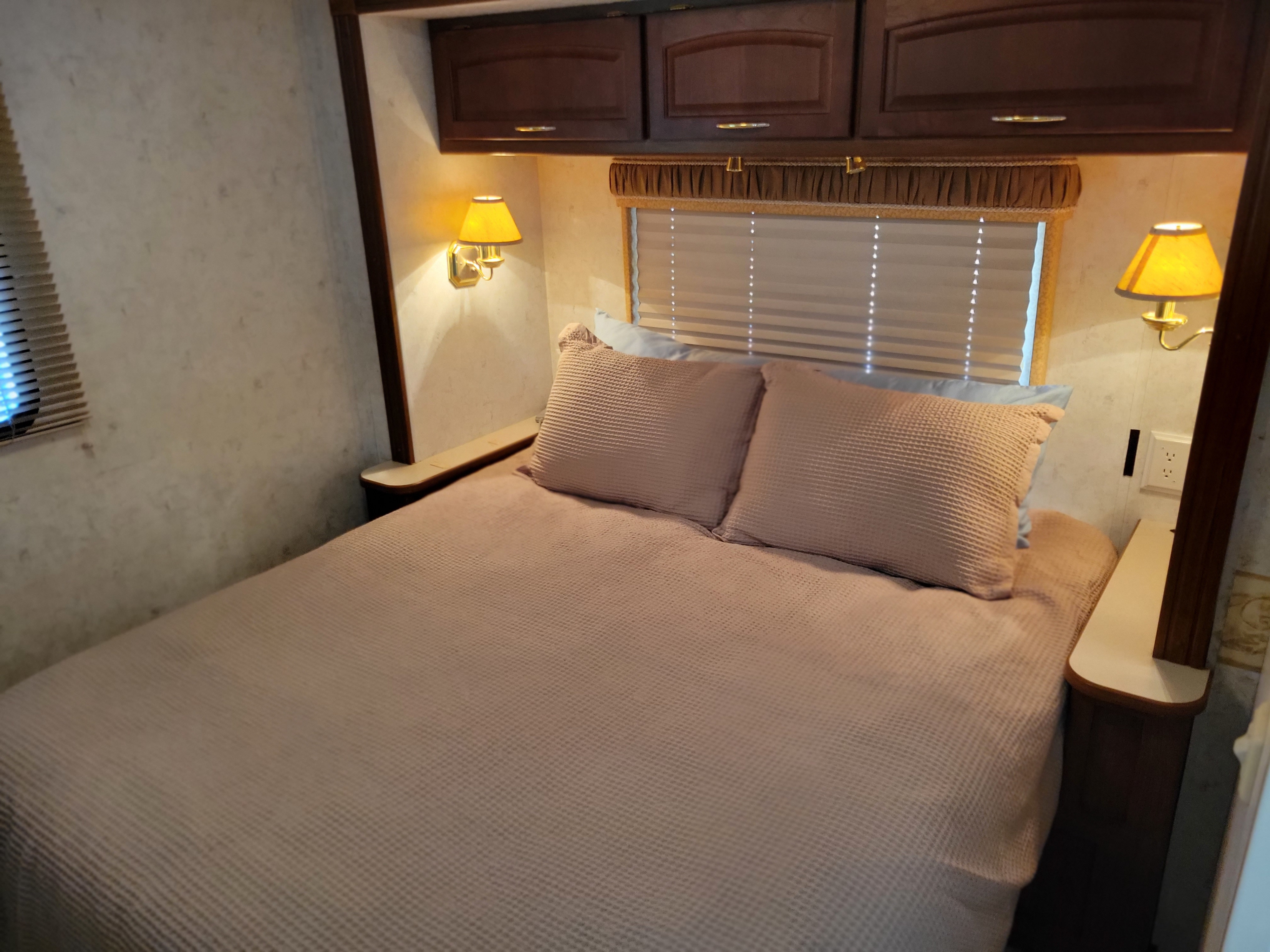 Cozy RV rental bedroom featuring a bed with textured blanket, warm wall lamps, window blinds, and convenient outlets.. Itasca Sundancer 2004