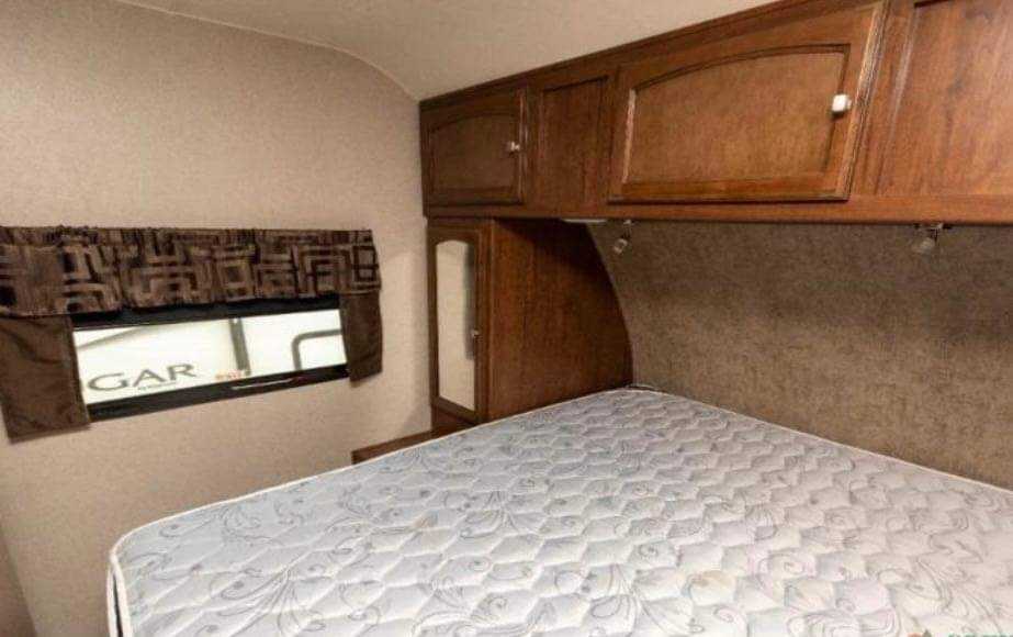 View of an RV rental bedroom: a white, quilted mattress, wooden overhead cabinets, and a window with brown valance.. Coachmen Other 2018