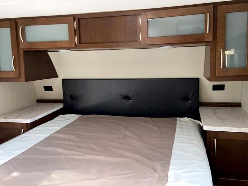 Comfortable RV rental bedroom features a bed with a black headboard, overhead cabinets, and side shelves.. Prime Time Crusader 2018