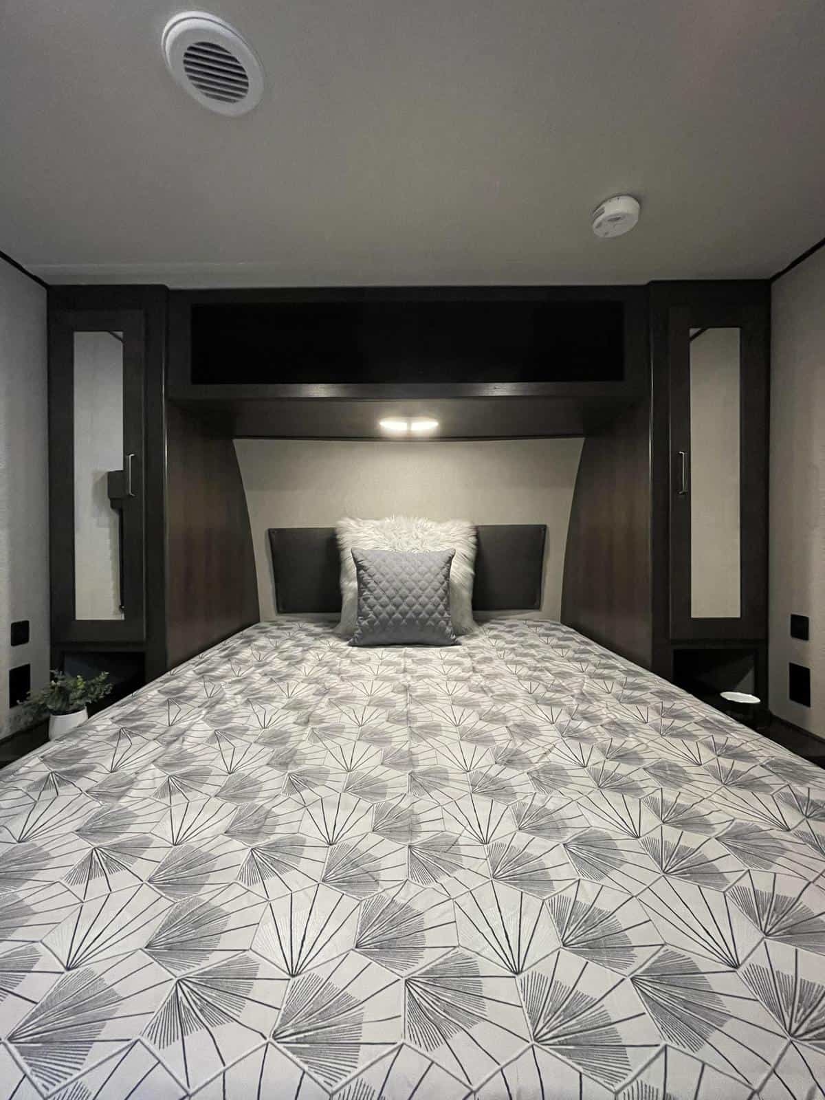 View of a cozy RV rental bedroom featuring a bed with a patterned comforter, overhead storage, and mirrored cabinets.. Grand Design Transcend Xplor 265BH 2022