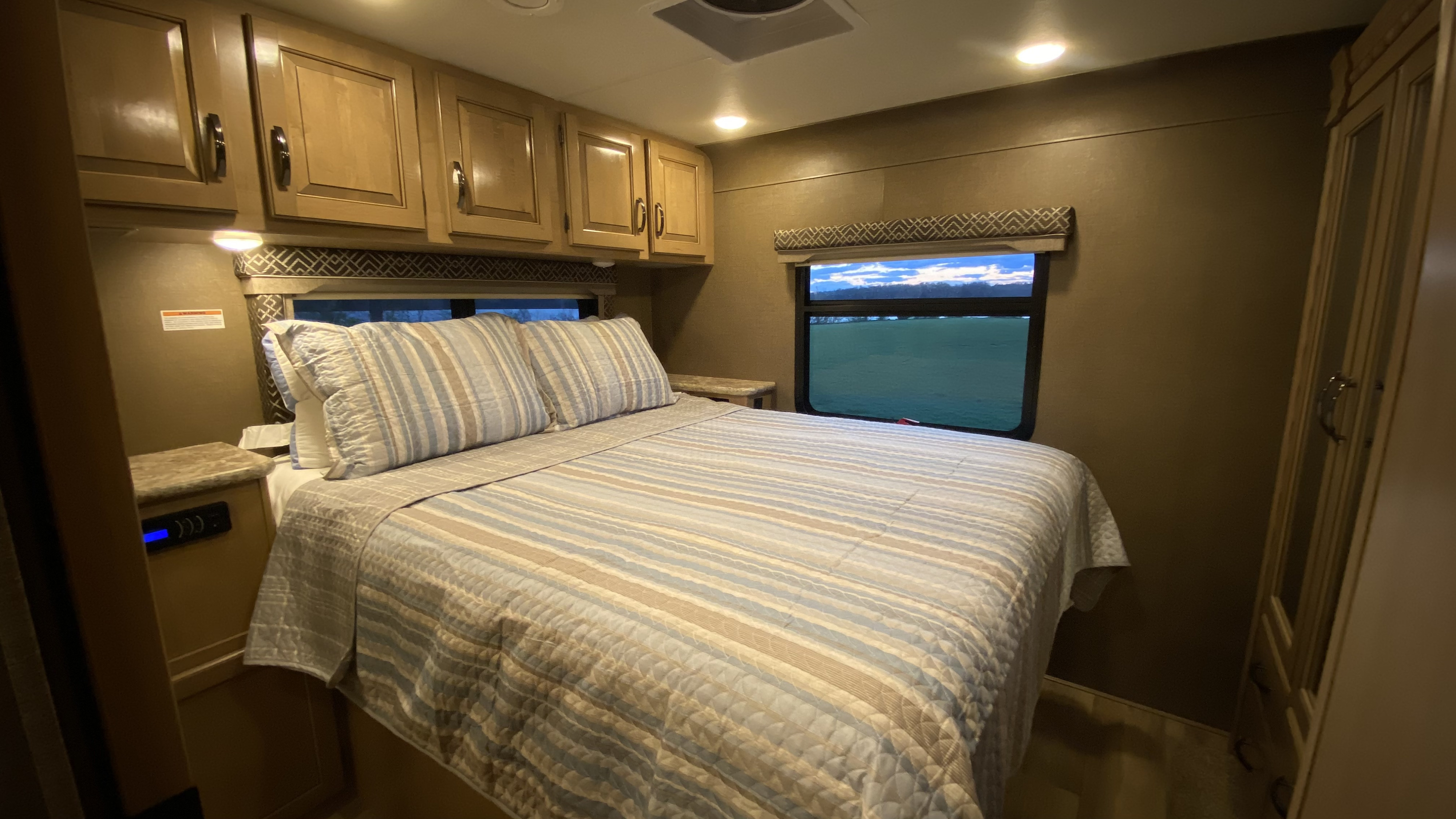 Comfortable RV rental bedroom featuring a striped bed, overhead cabinets, and a window with a pleasant outdoor view.. Thor Four Winds 2018
