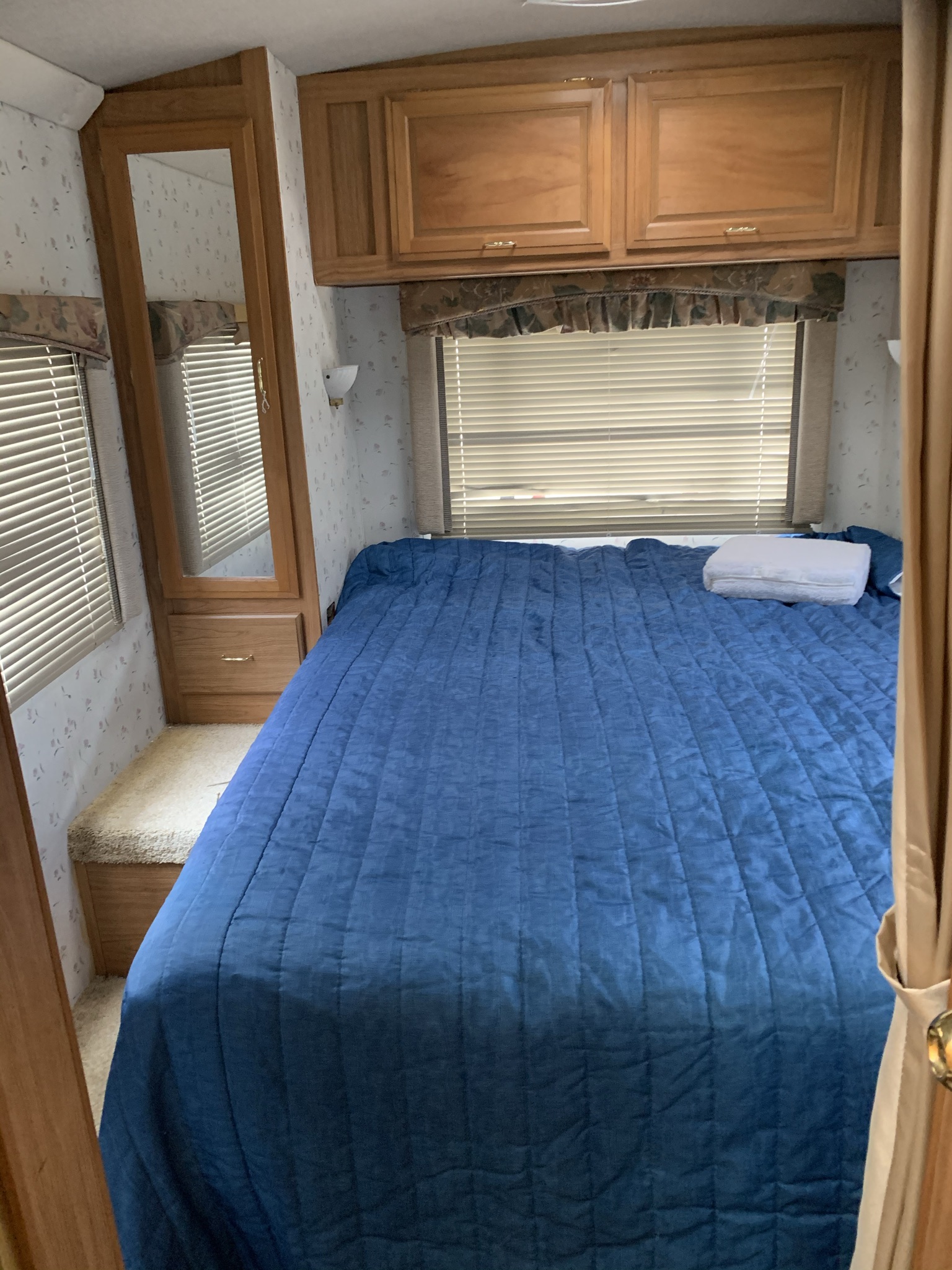 Experience comfort in this RV rental bedroom featuring a blue bed, wooden cabinets, windows with blinds, and a tall mirror.. Ford Tioga 1998