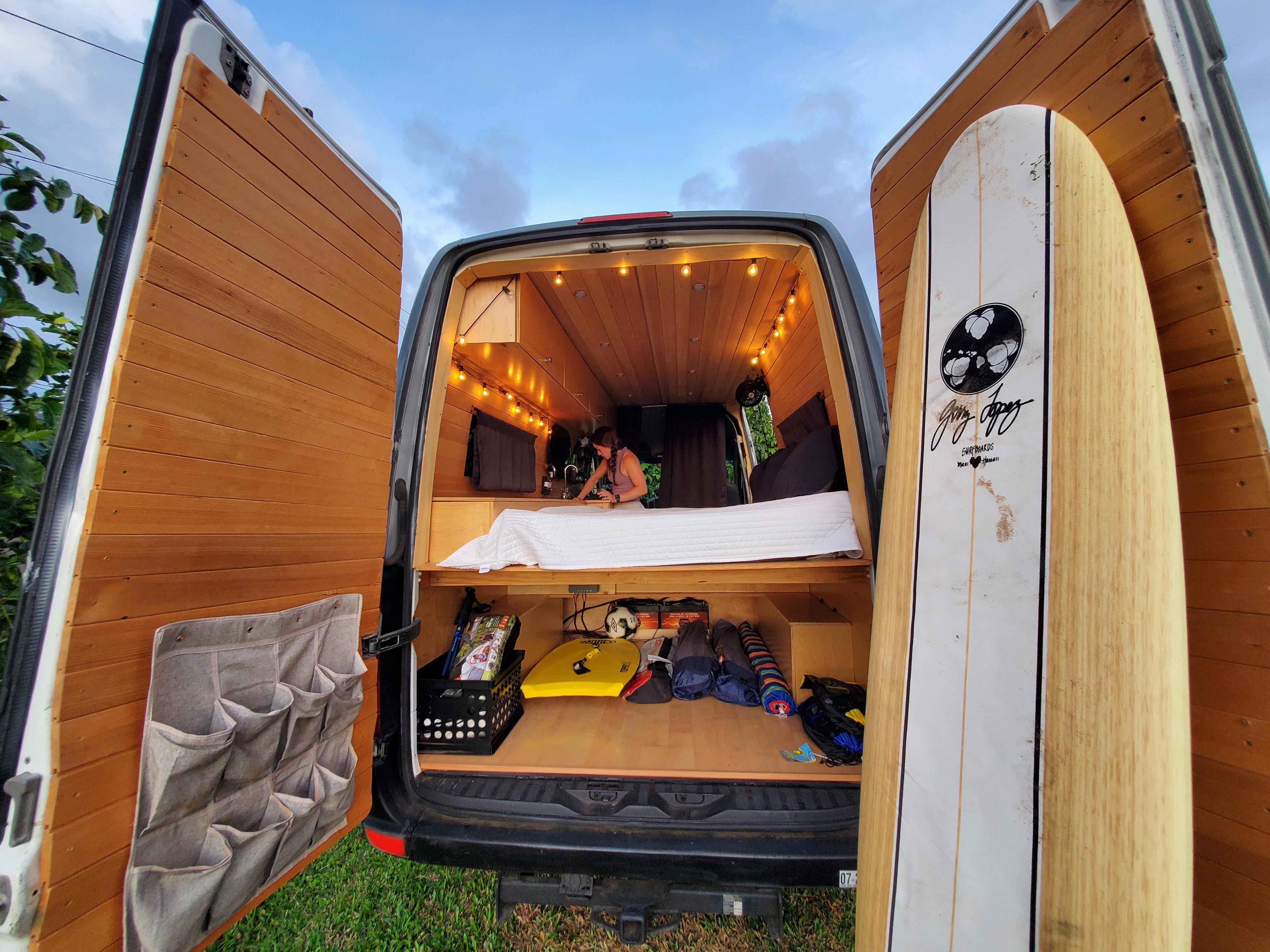 Rear of an RV rental van with doors open, showcasing a cozy, wood-paneled interior with a bed, warm lights, and gear, including a surfboard.. Mercedes-Benz Sprinter 2007