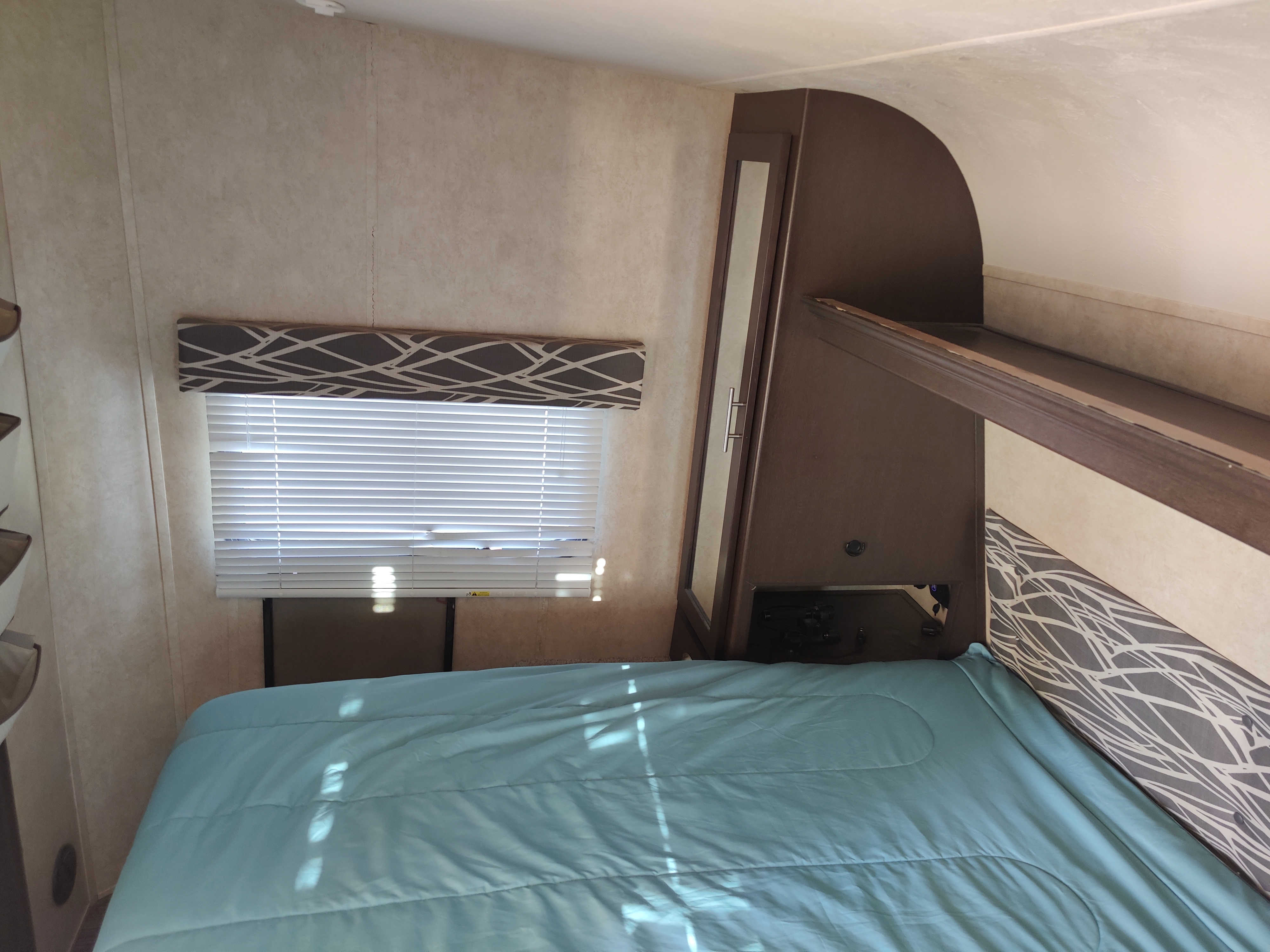 Cozy RV rental bedroom with a light blue bed, a window with blinds, and built-in storage, illuminated by sunlight.. Forest River Stealth 2015