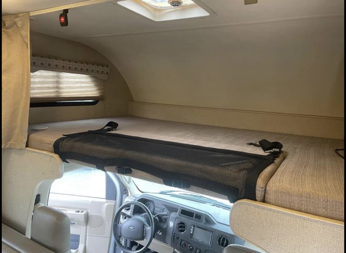 Interior of an RV rental: The cab-over bunk bed with safety net, a window with blinds, and the vehicle's dashboard.. Thor Motor Coach Four Winds 2019