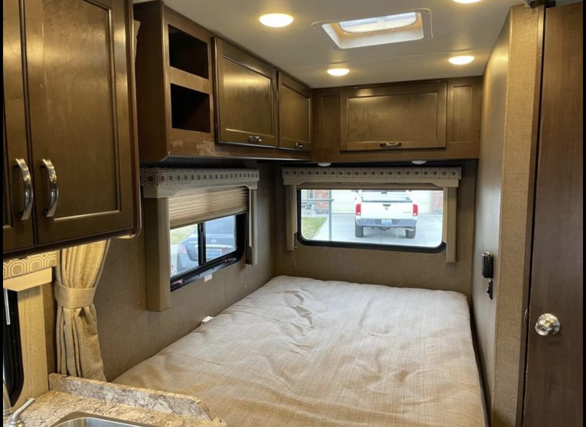 This RV rental's bedroom includes a bed, overhead cabinets, windows showing an outside view, and ceiling lights.. Thor Motor Coach Four Winds 2019