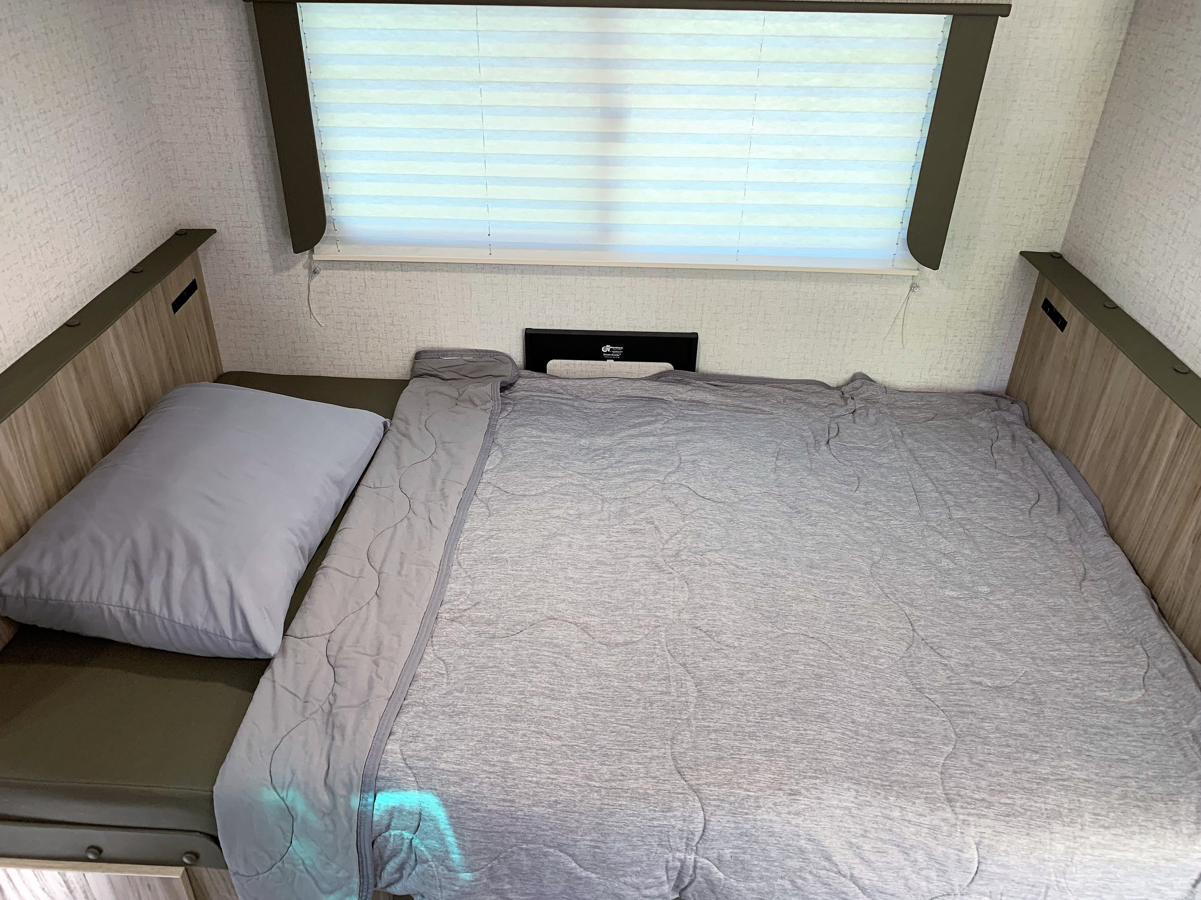 Cozy RV rental bedroom featuring a neatly made bed with a grey comforter and pillow, beneath striped window blinds.. Winnebago Minnie Winnie 2022