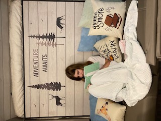 Inside an RV rental: A girl reads comfortably on a bed surrounded by adventure-themed pillows and decor.. Keystone RV Passport SL 2021