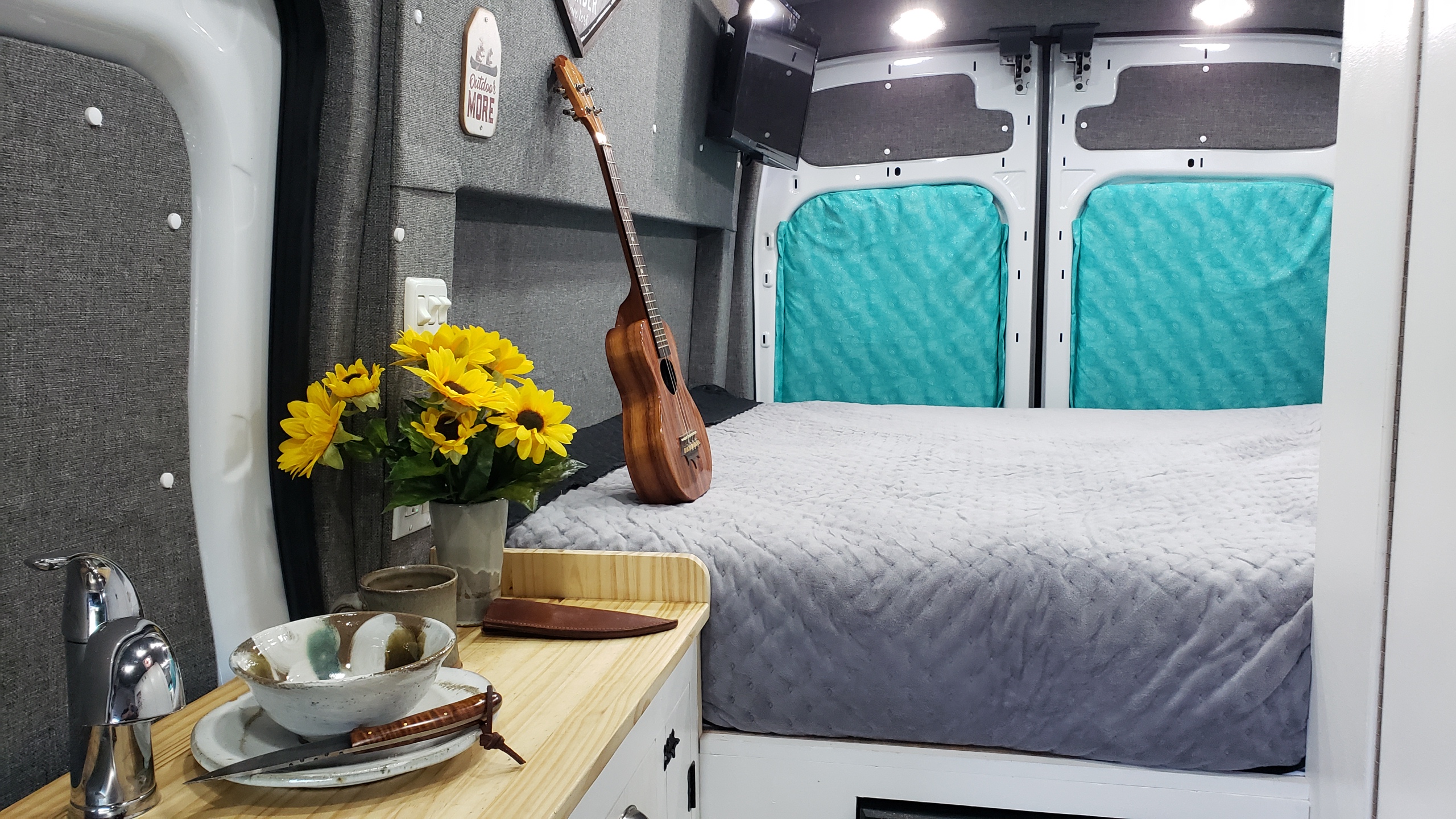 Cozy RV rental interior showing a bed, kitchen counter with a sink, and a ukulele, perfect for travel.. Ford Transit Custom 2021